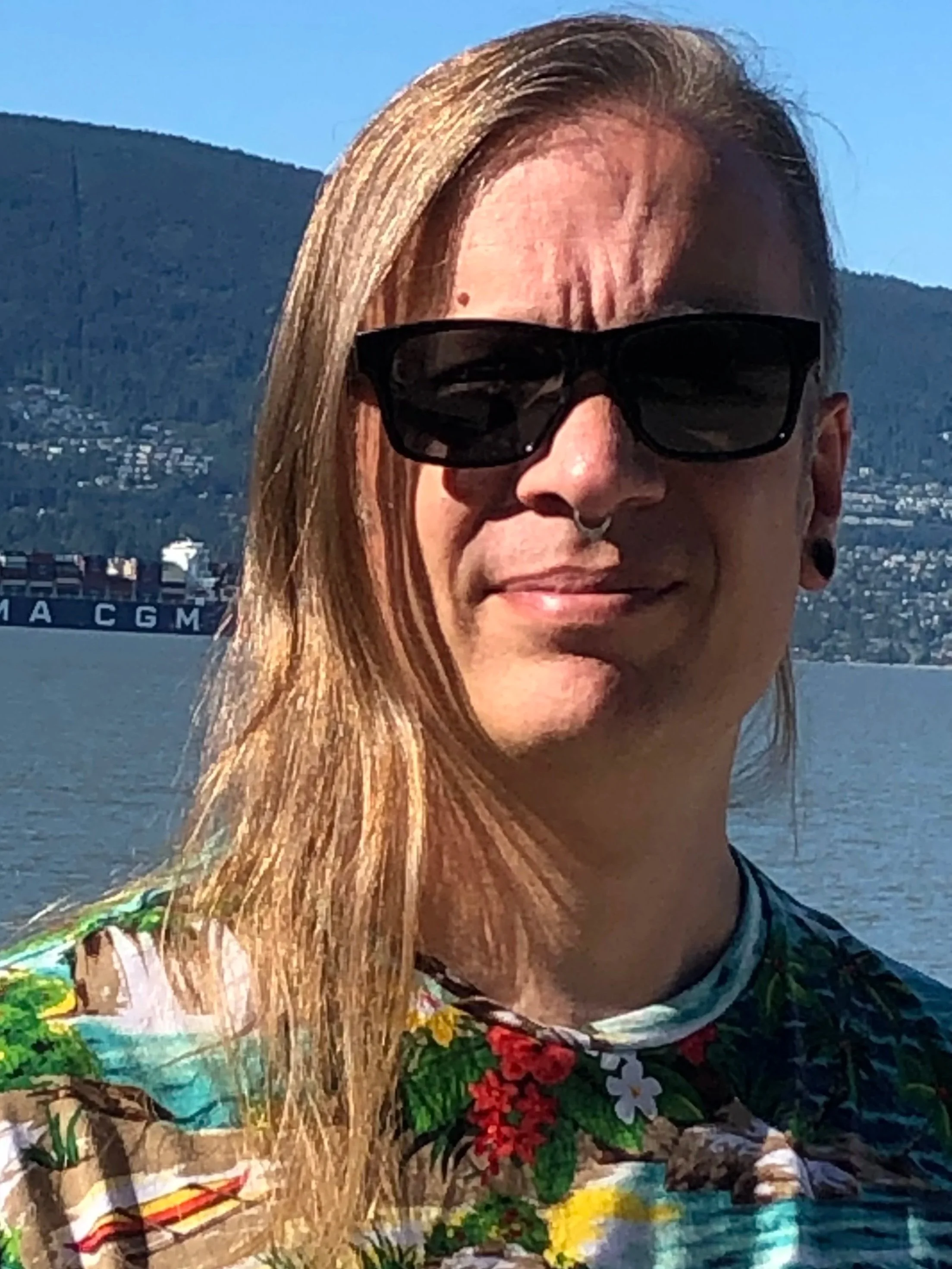 A person with long blonde hair wearing black sunglasses and a colorful patterned shirt, standing outdoors near a body of water with a mountain in the background.