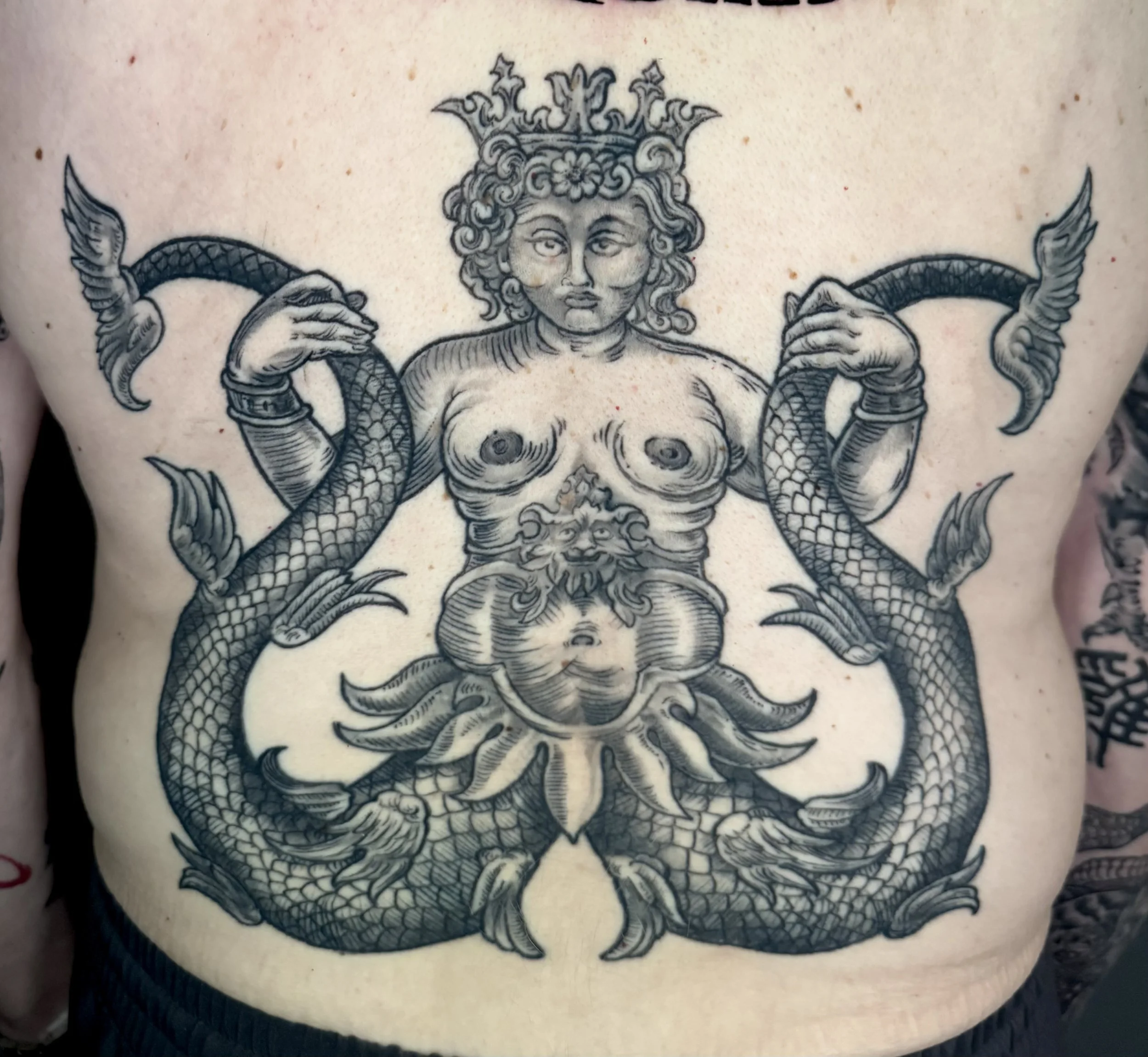 Tattoo of a mythological female figure with a crown, holding two serpents, with a lion's face on her chest, and a decorative floral element below.