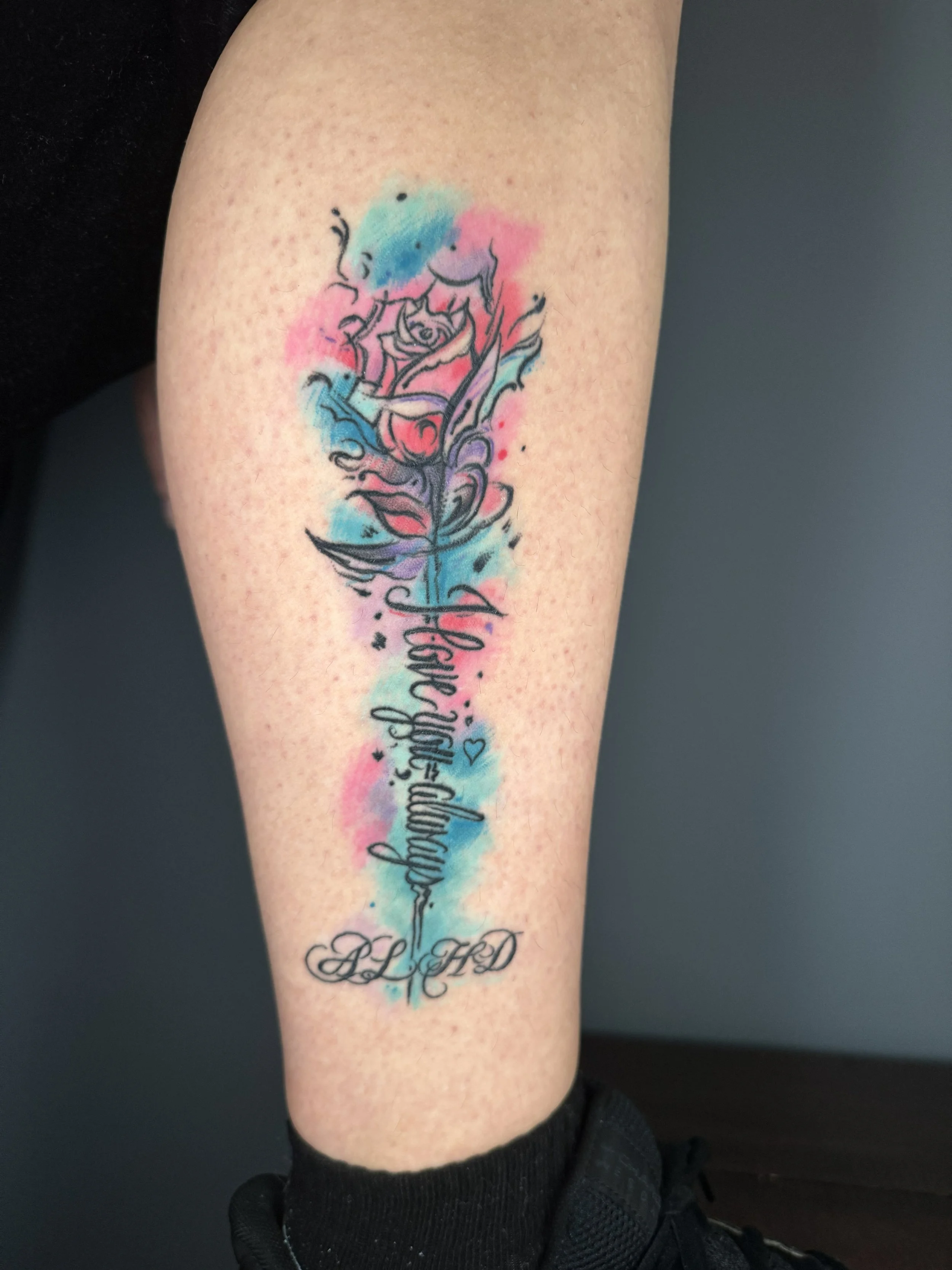 A tattoo on a person's arm featuring a watercolor-style pink, blue, and purple background with black script text and a small heart detail.