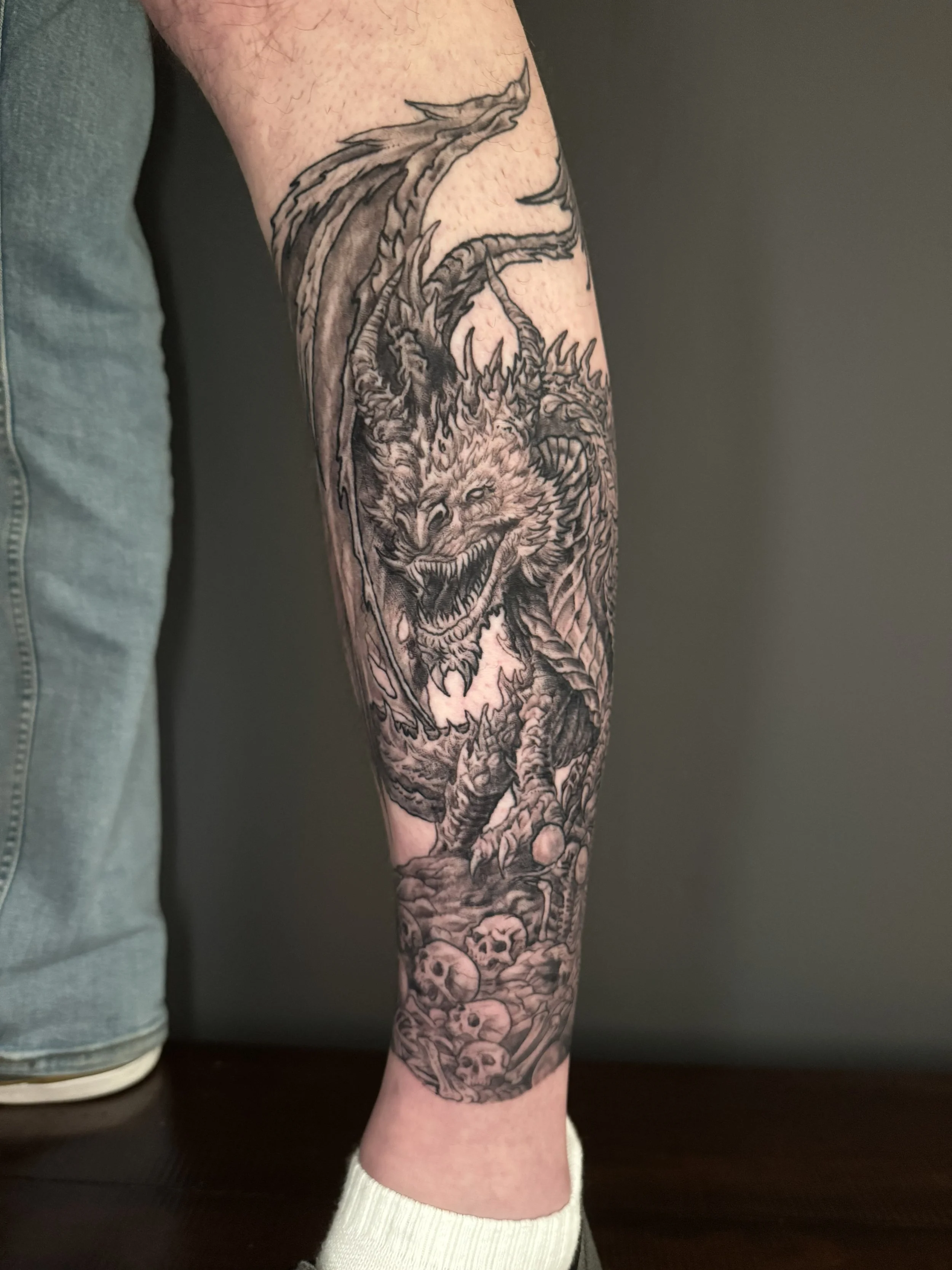 Black and gray tattoo of a dragon with a fierce expression, long body, and skulls on the lower part of the arm.