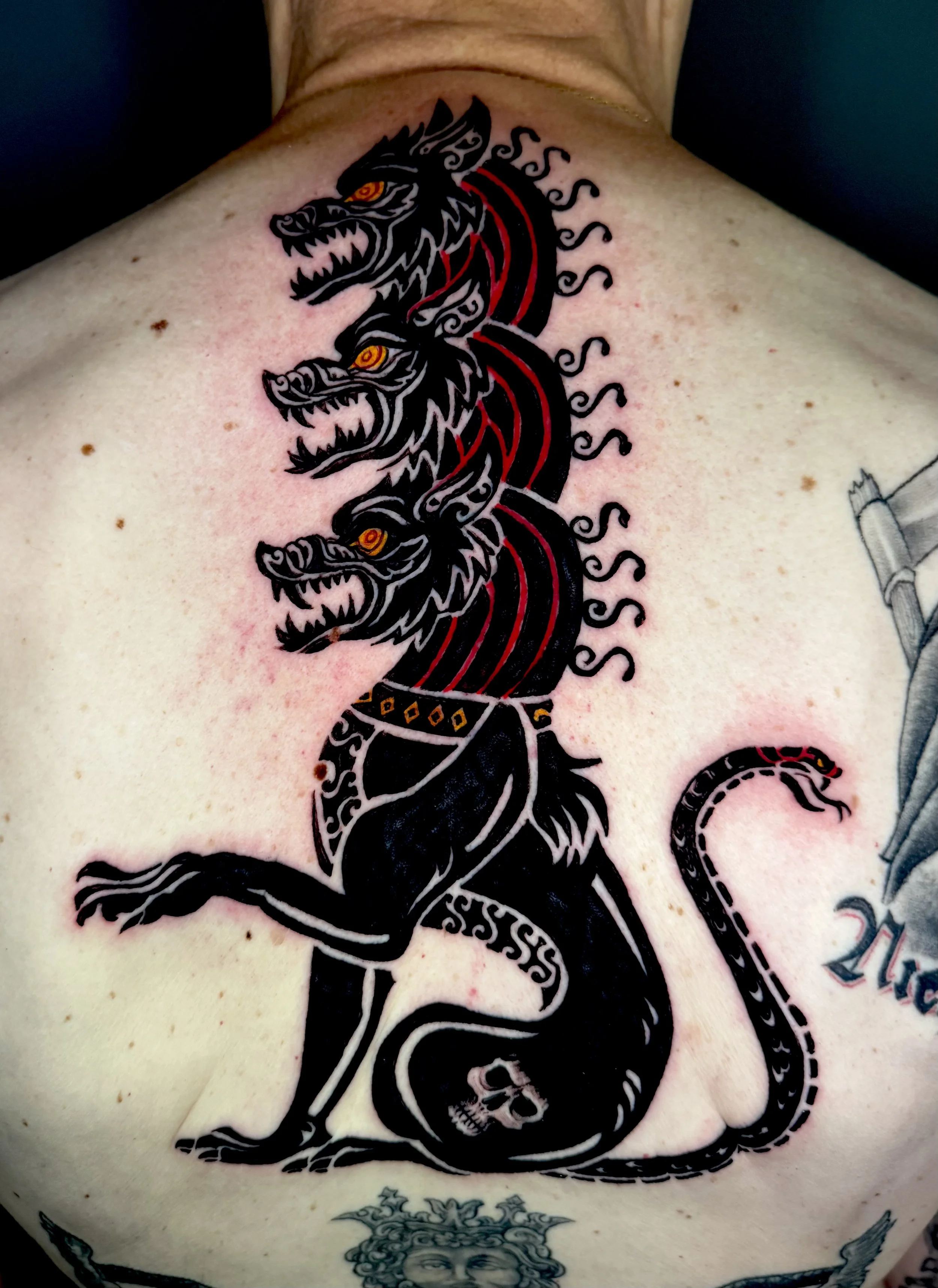 Tattoo of a black three-headed dog with orange eyes, sitting on a person’s chest, with a skull at its paw and a decorative collar.