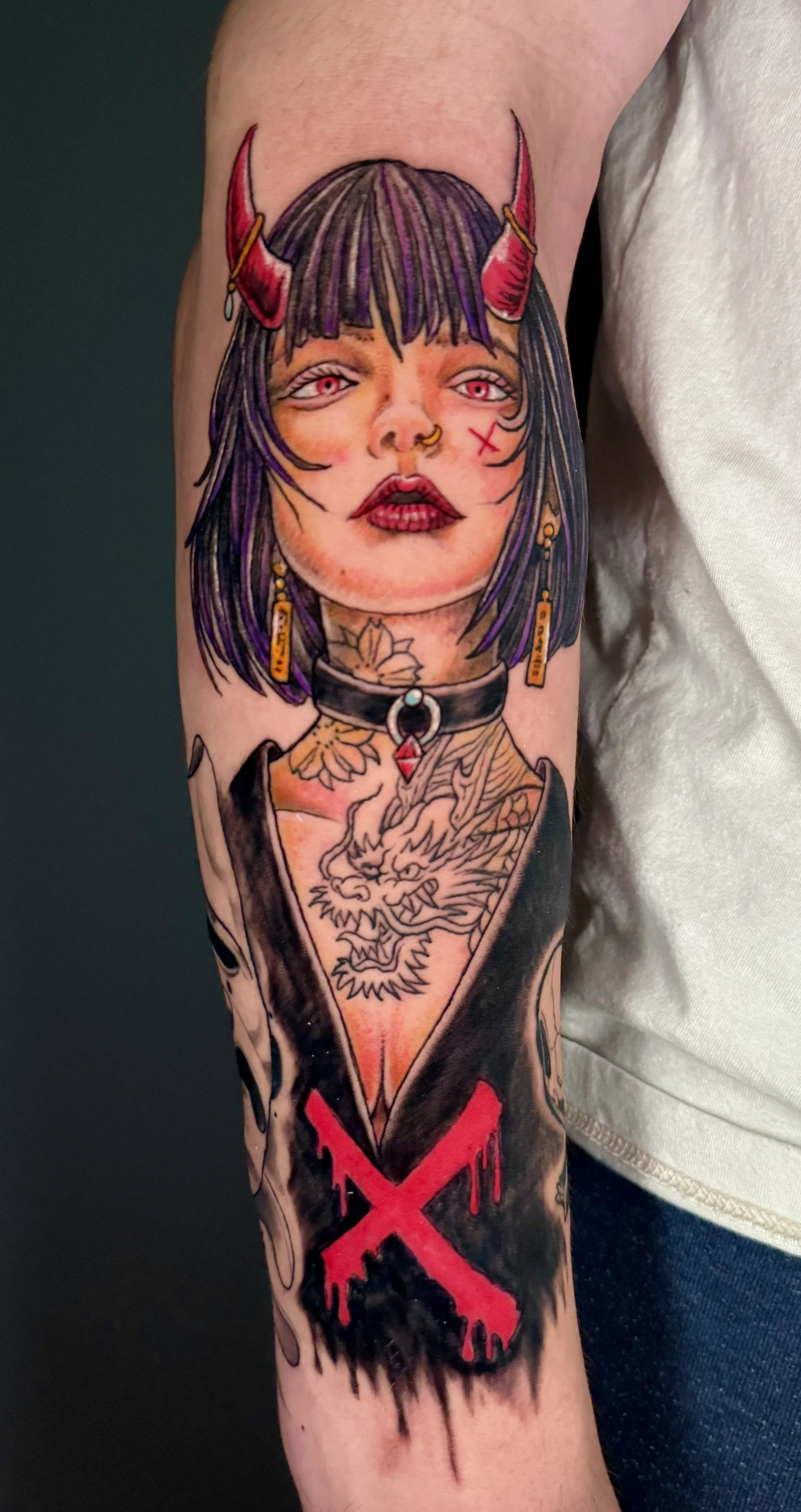 A colorful tattoo of a woman with black hair, purple streaks, red horns, and a choker with a dragon design on the arm.