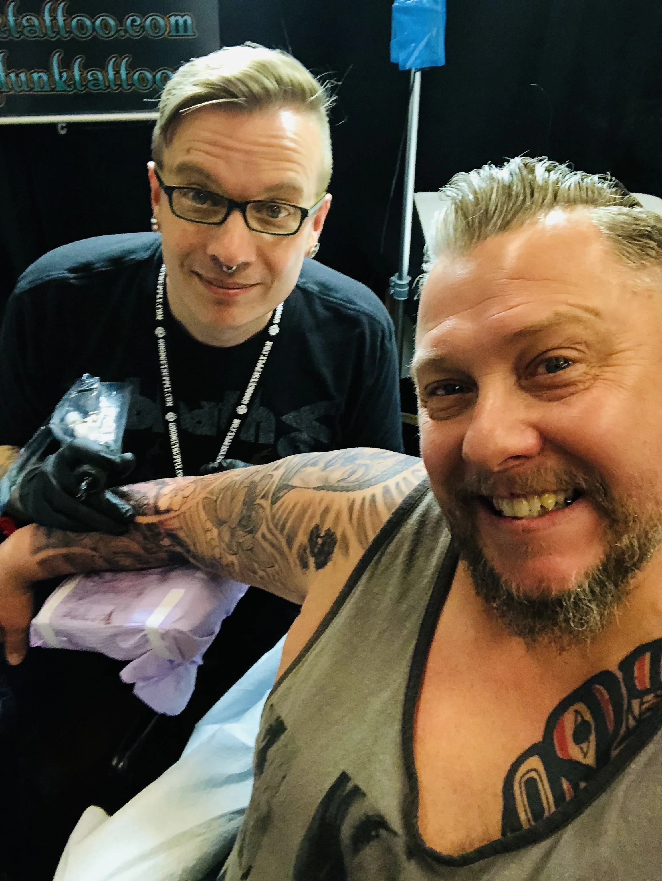 Two men smiling, one getting a tattoo on his arm by a tattoo artist. The tattoo artist has glasses, a septum piercing, and tattoos on his arm. The man receiving the tattoo has a beard and is wearing a sleeveless shirt. The background shows part of a sign and some equipment.
