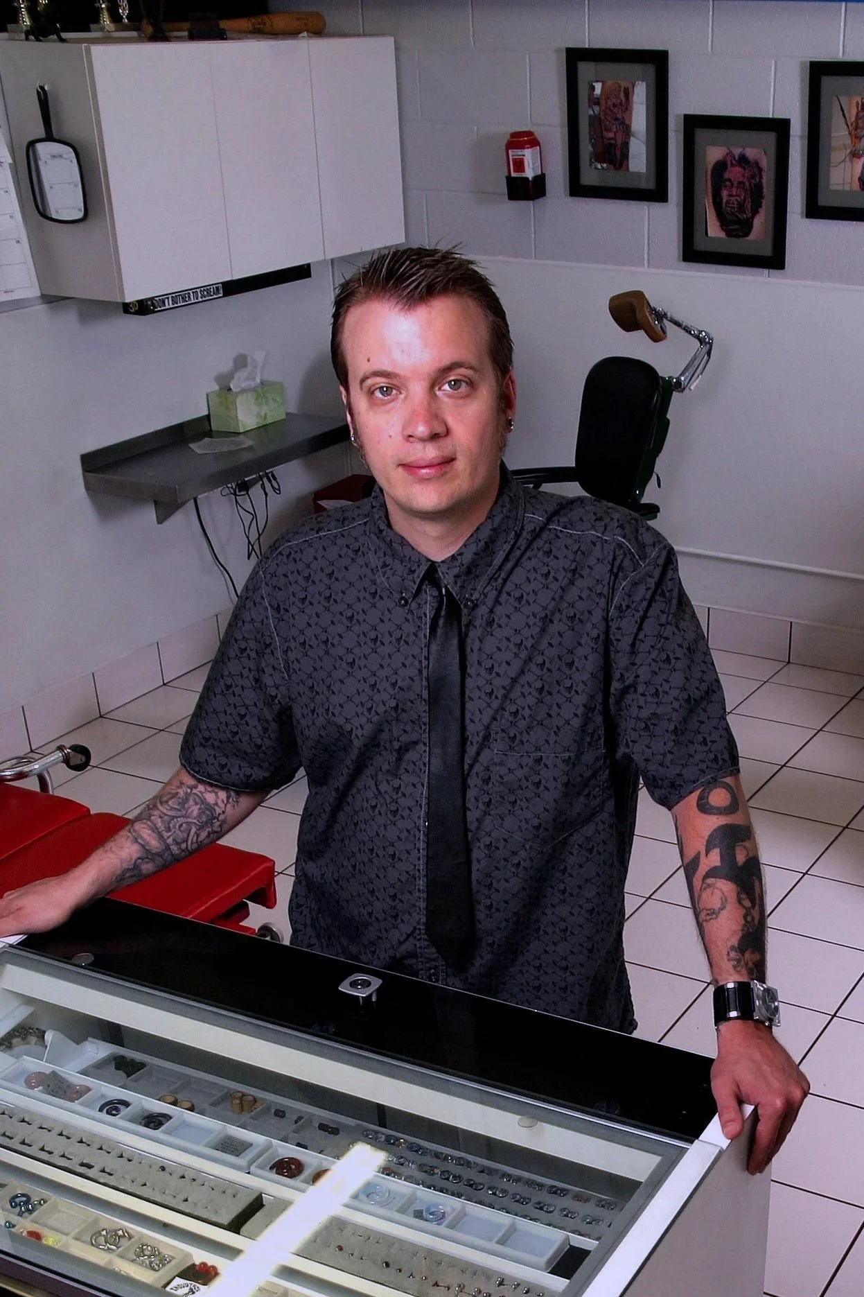 A man in a dark patterned shirt and black tie with tattoos on his arms is standing behind a glass display case filled with jewelry and watches in a retail store. He has short hair and is looking at the camera.