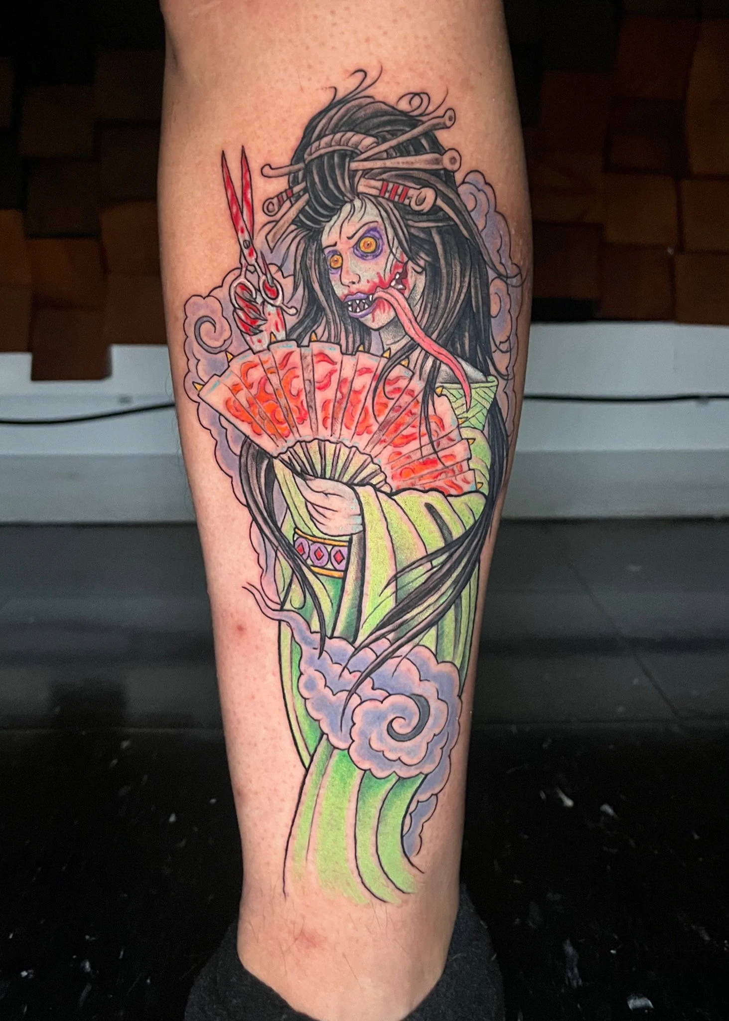 Tattoo of a scary female character holding a fan, with long black hair and a menacing expression, surrounded by smoke.