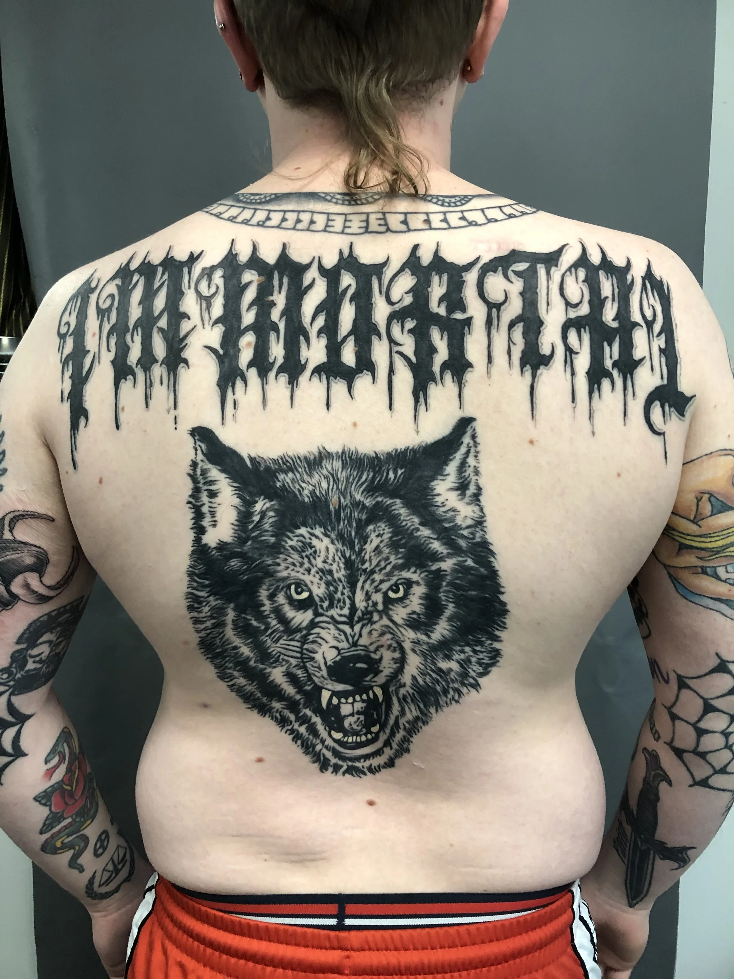 A person with short hair and tattoos on their arms and back, displaying a large tattoo of a snarling wolf's head in black ink on their chest. The tattoo is detailed and realistic, with the wolf's teeth bared and eyes intense. Above the wolf tattoo, t