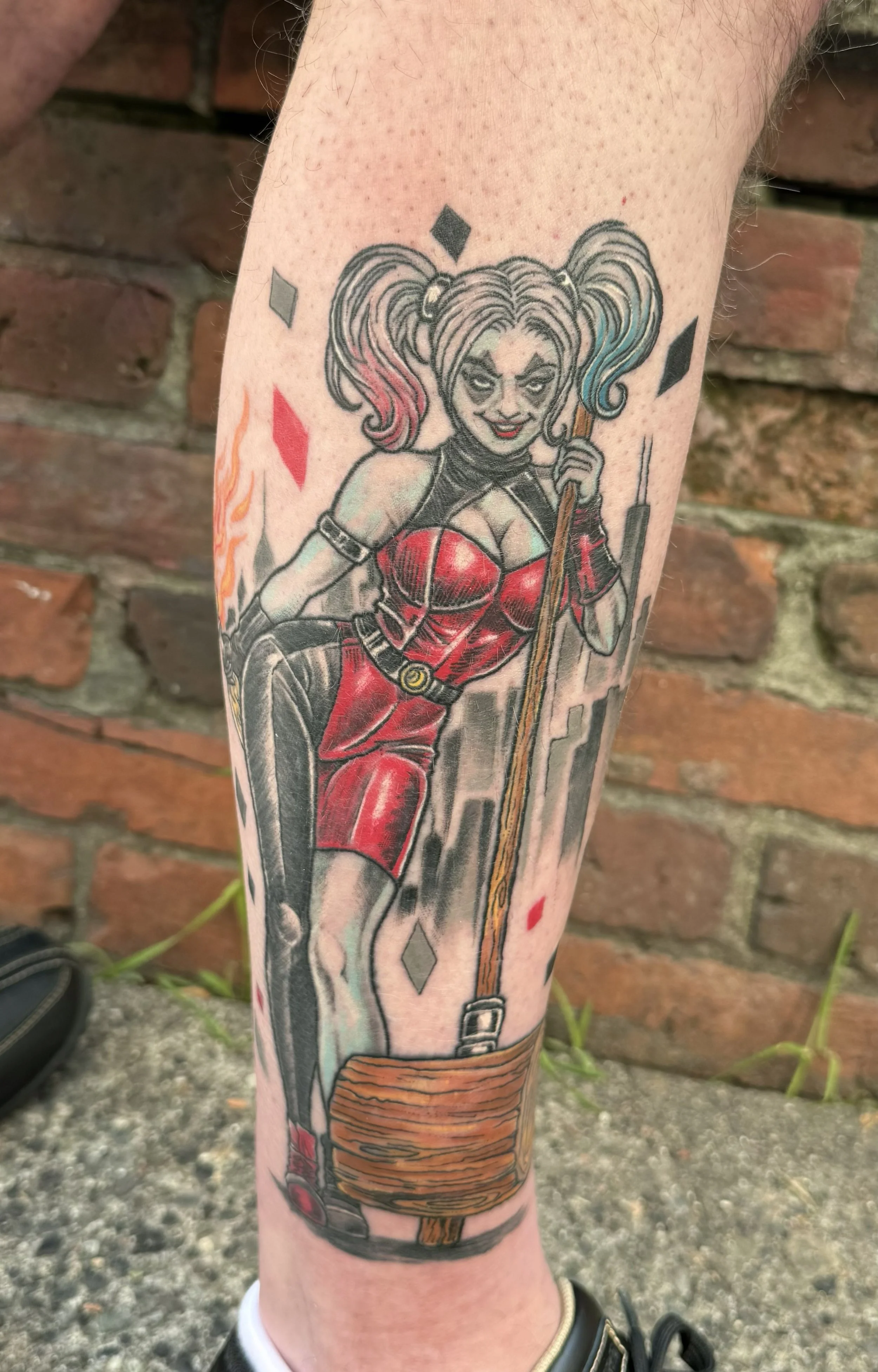 Tattoo of Harley Quinn holding a baseball bat, wearing a red and black outfit with puffy hair dyed pink and blue, with playing card symbols around her.