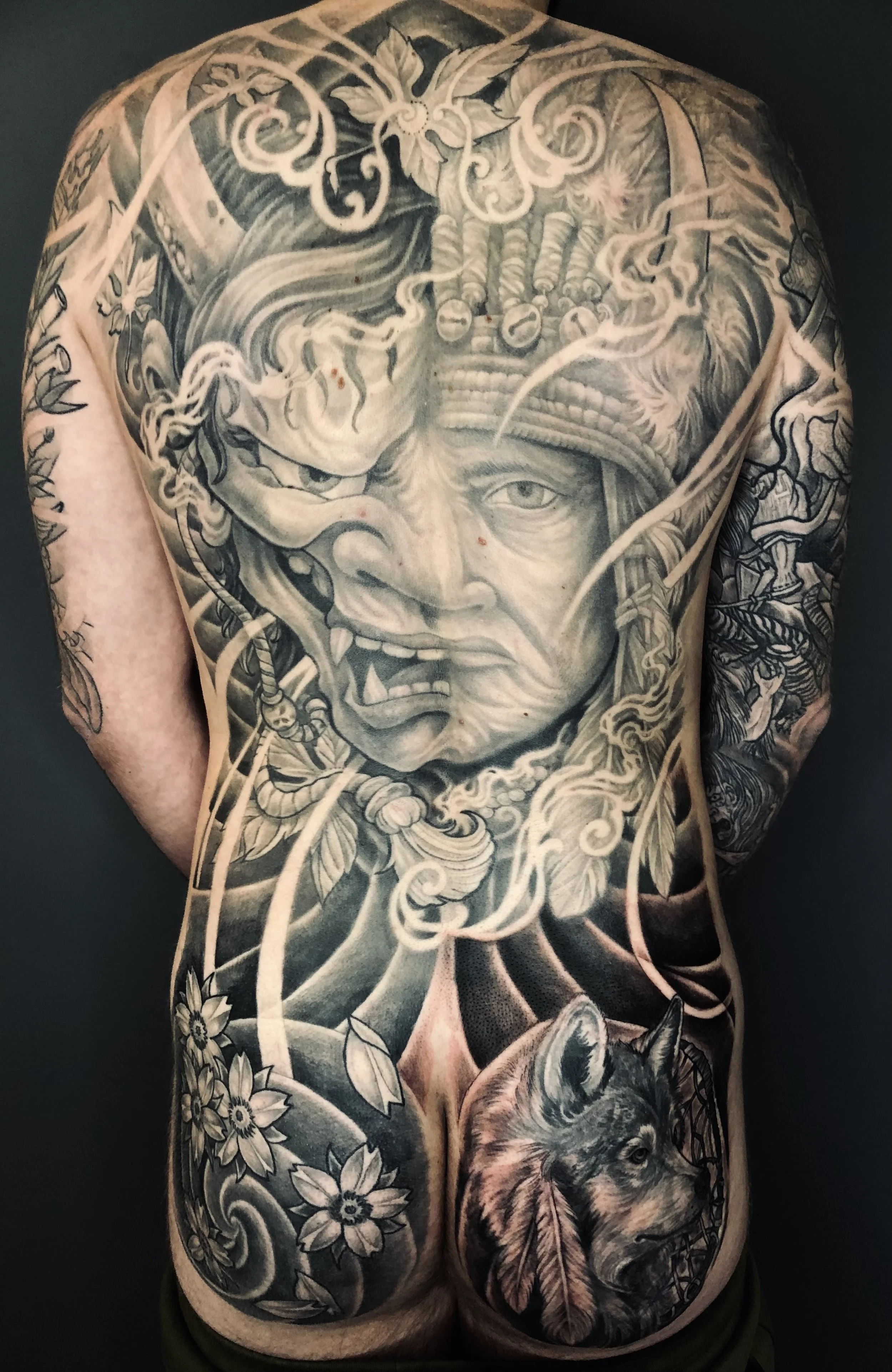 Black and gray tattoo covering a person's back, depicting a grotesque face with a crown of thorns and a wolf's head with a leaf.