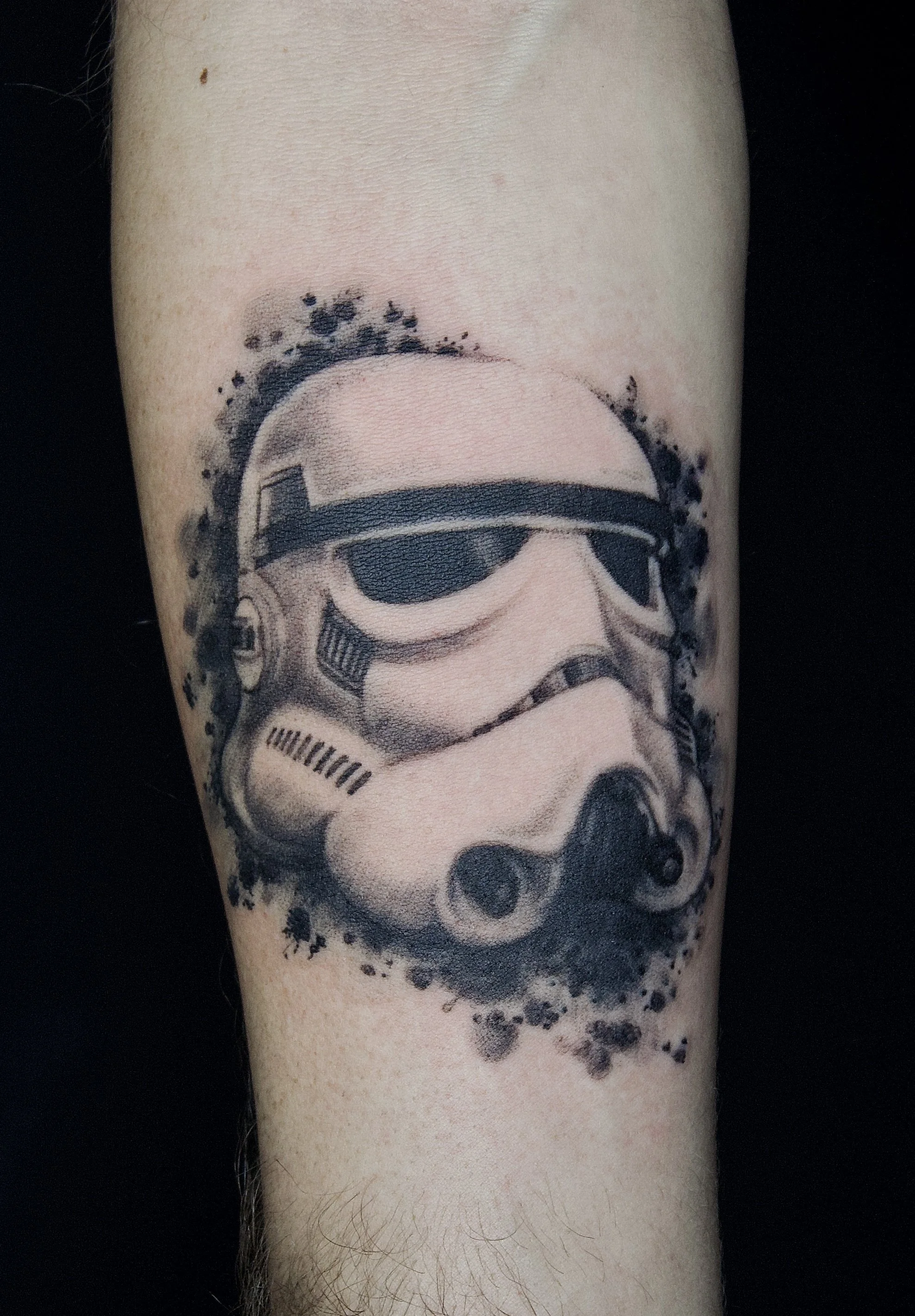 Tattoo of a Stormtrooper helmet from Star Wars, in black and gray ink, with a watercolor splatter background.