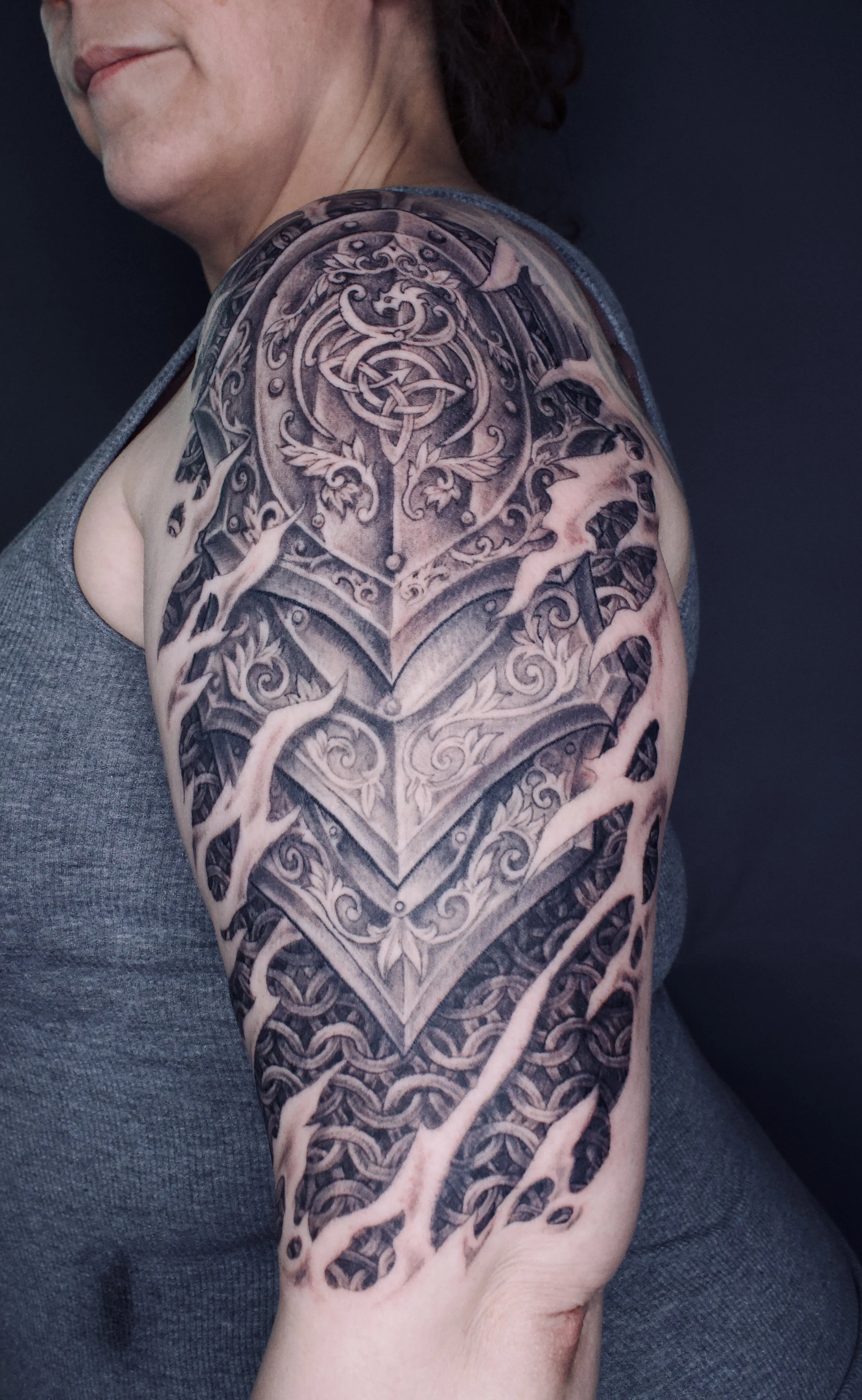 Close-up of a person's upper arm and shoulder with a detailed black and gray tattoo featuring intricate designs and patterns.