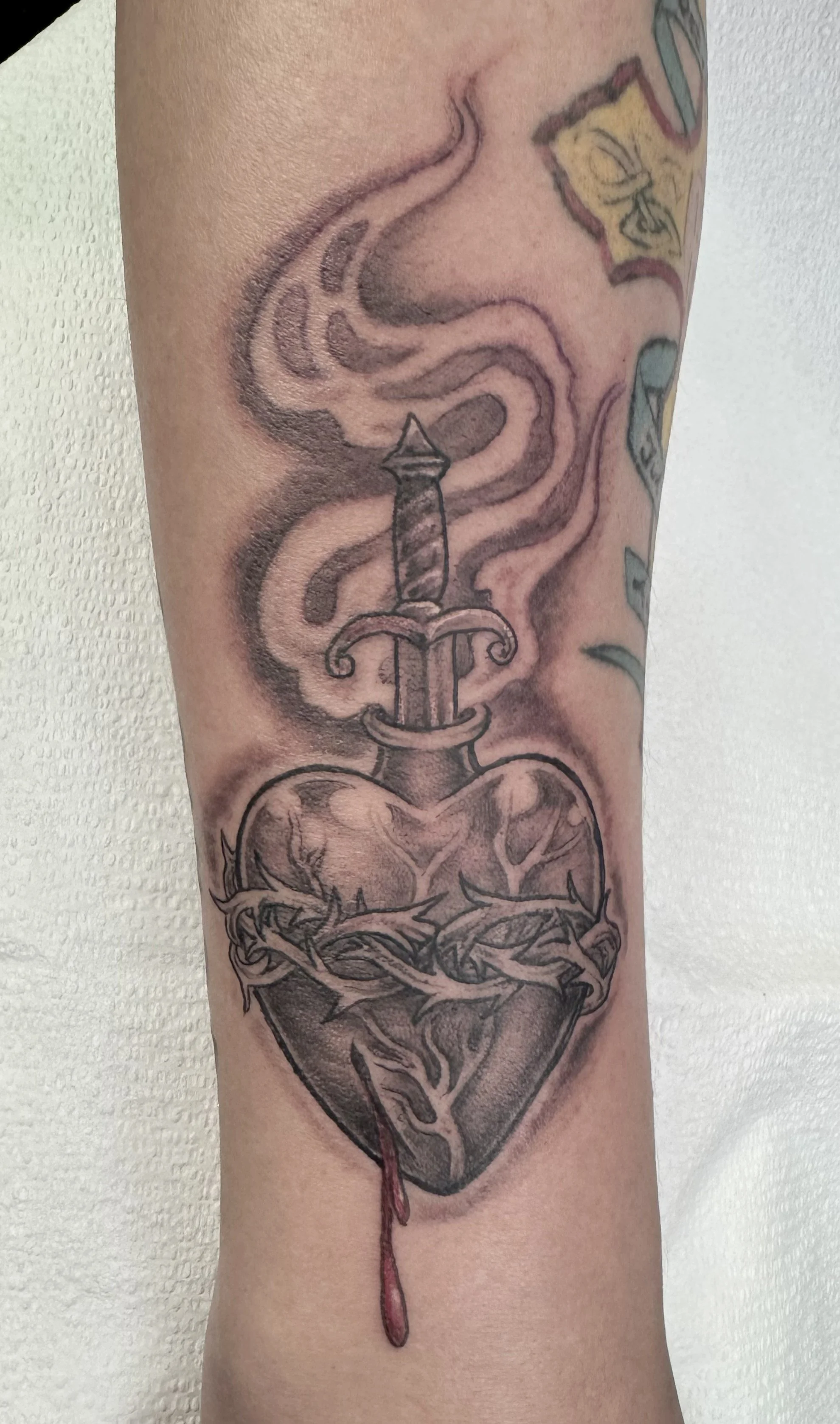 A tattoo of a dagger with a heart and a crown of thorns, with smoke swirling around it.