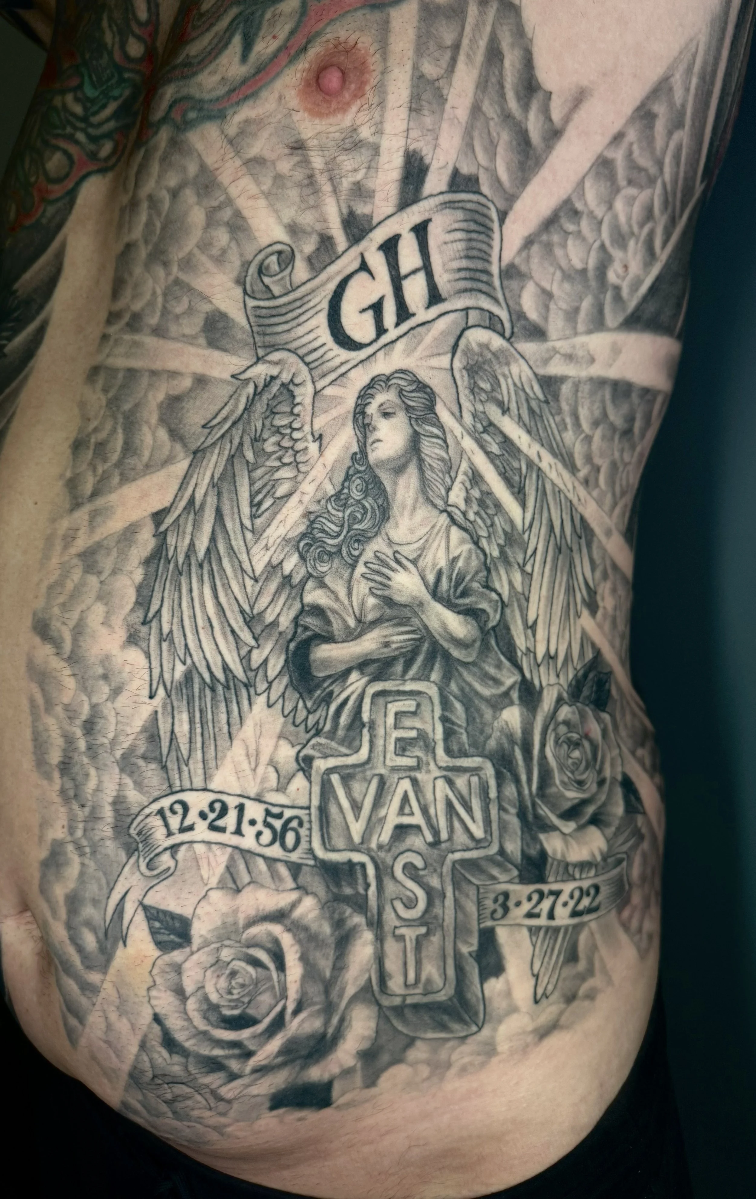A detailed black and gray tattoo on a person's chest and stomach, featuring an angel with wings, a banner with the initials 'GHL,' roses, a cross with the name 'VAN,' and dates including '12-21-56' and '3-27-22.'