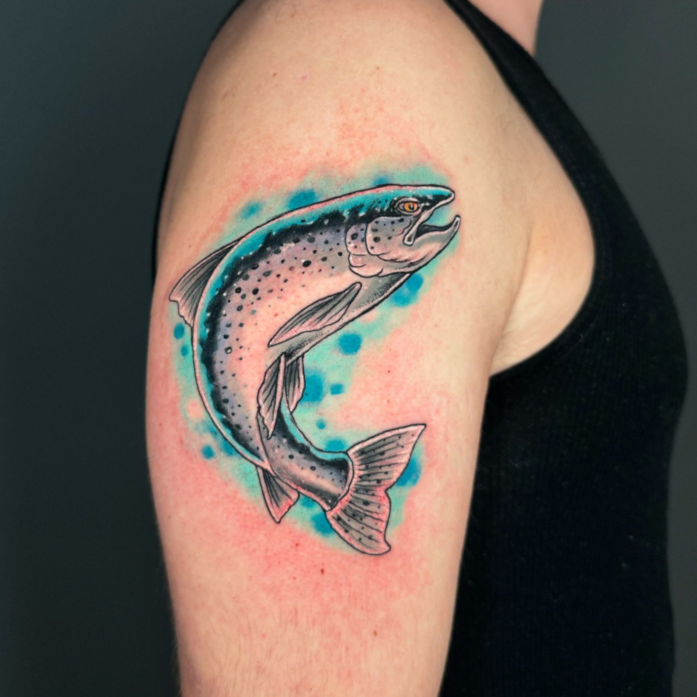 Colorful tattoo of a salmon fish on person's upper arm, with blue and pink watercolor background effects.