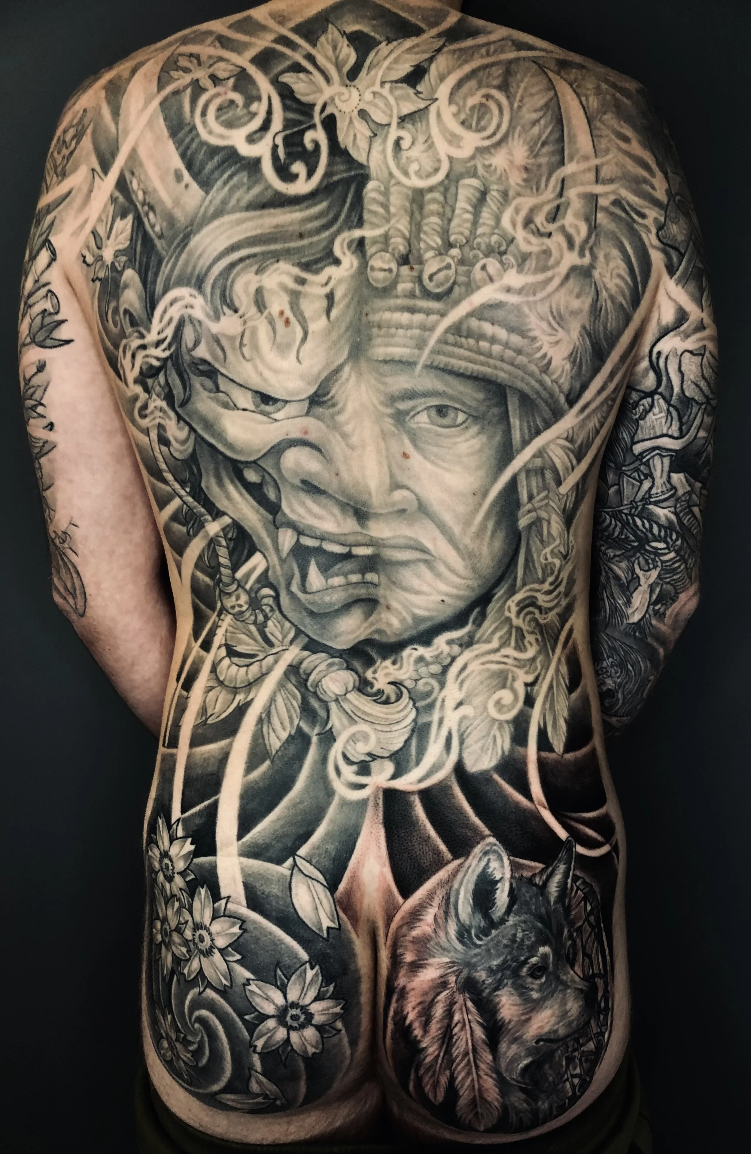 Black and gray tattoo covering the back and buttocks, featuring a fierce face with fierce expression, a wolf's head, flowers, leaves, and swirling patterns.