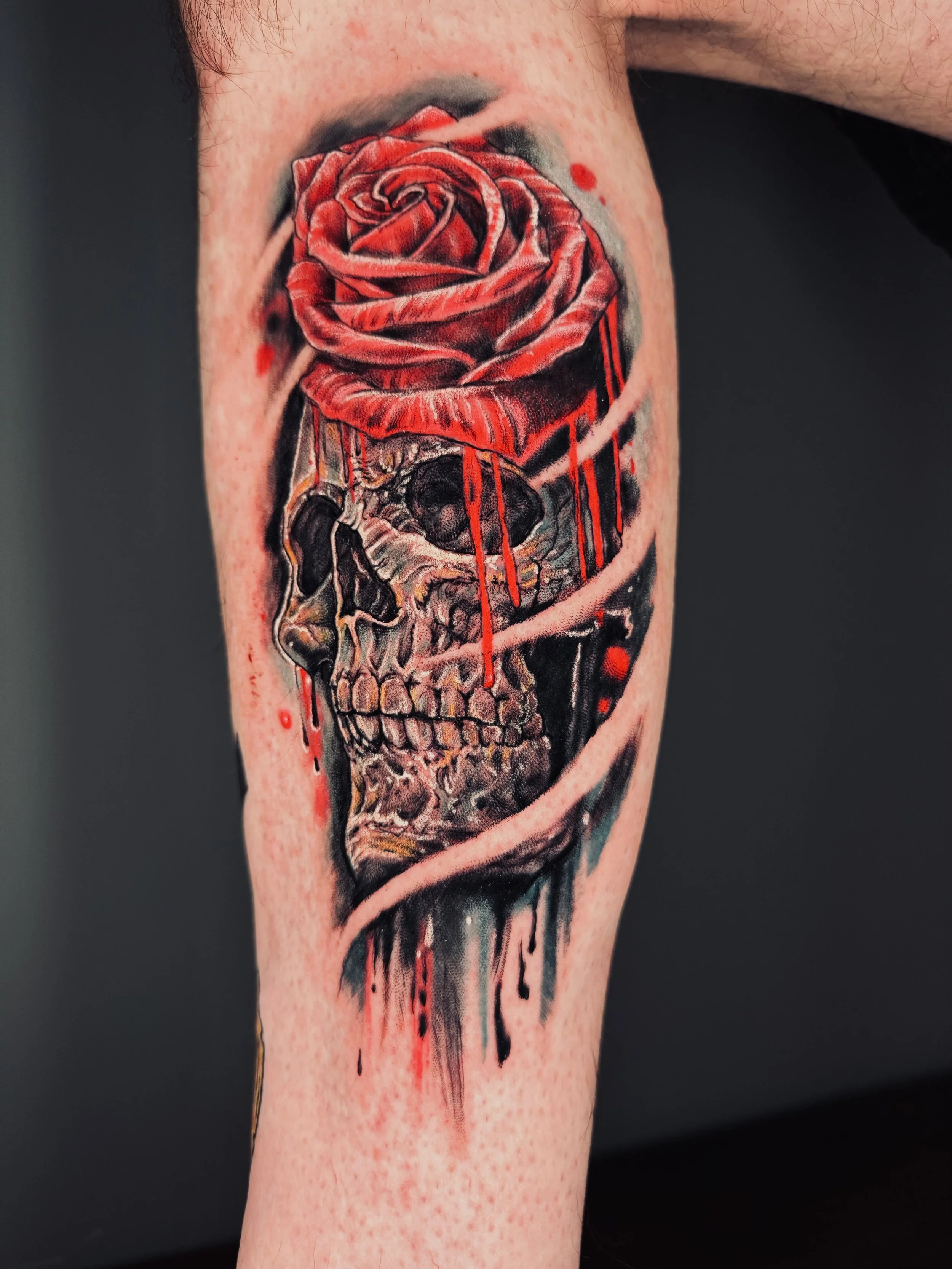 Tattoo of a skull with a red rose on top, with red and black ink accents and a pink swirl surrounding the skull.