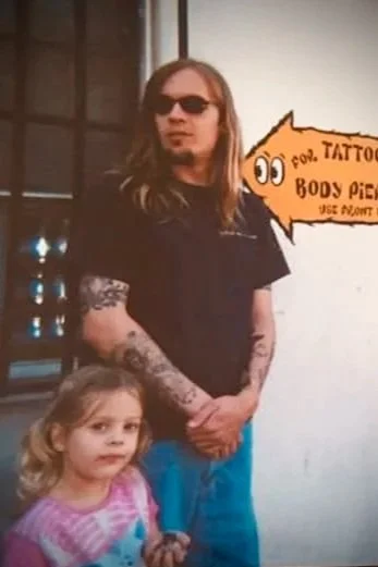 A man with long hair, sunglasses, and tattoos on his arm standing next to a young girl with blonde hair, in front of a wall with a tattoo shop sign.