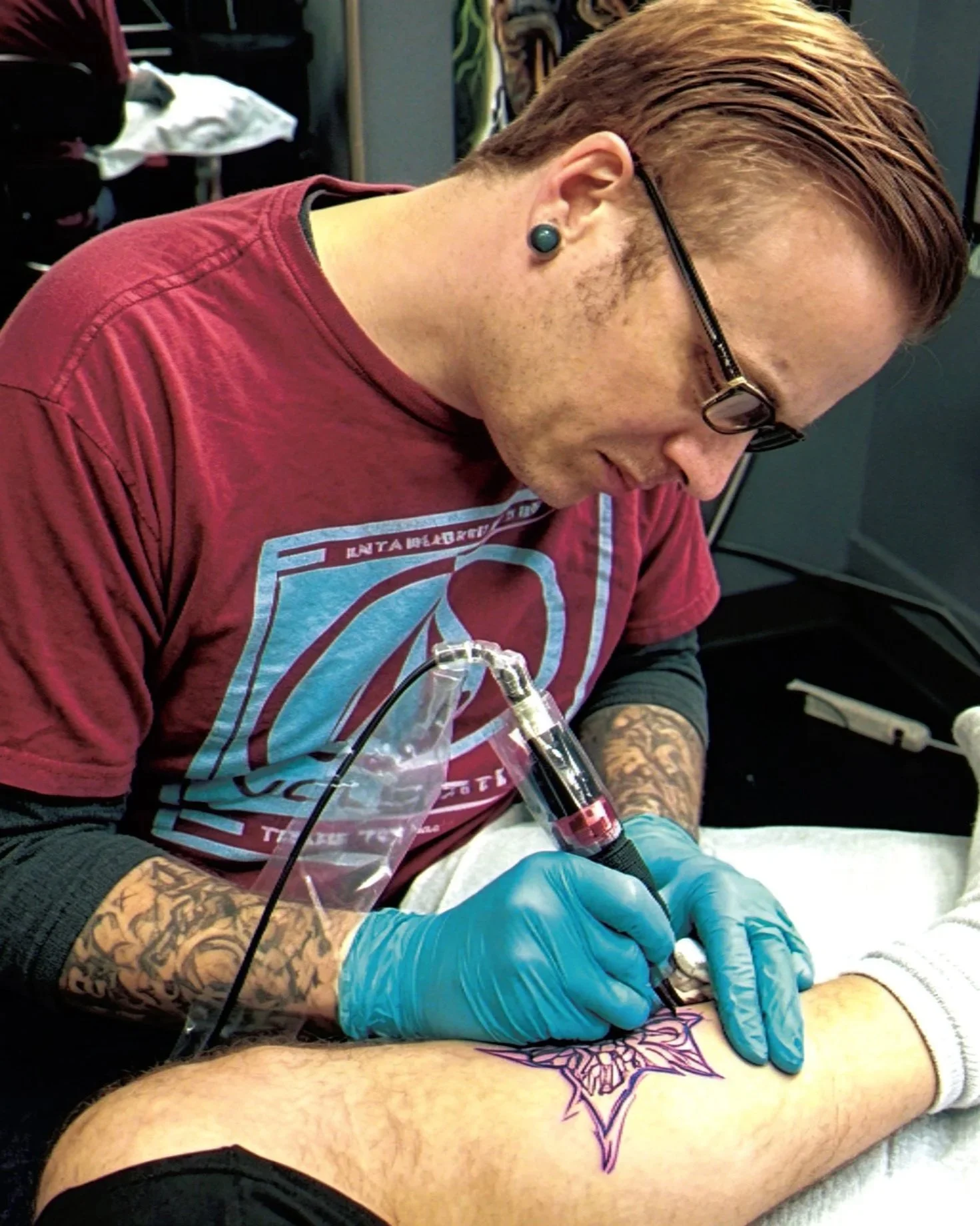 A man with glasses and tattoos on his arms is tattooing a design on another person's leg. He is wearing blue gloves and a smartwatch.
