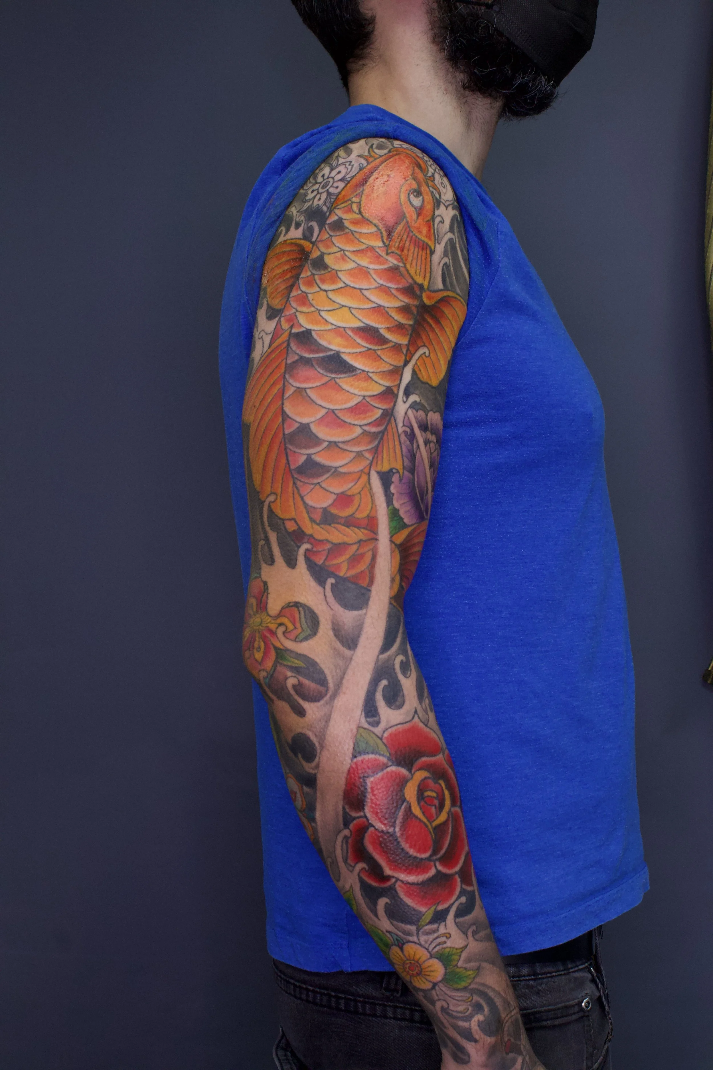 A person with a large colorful koi fish tattoo on their arm, wearing a blue sleeveless shirt, with a dark background.
