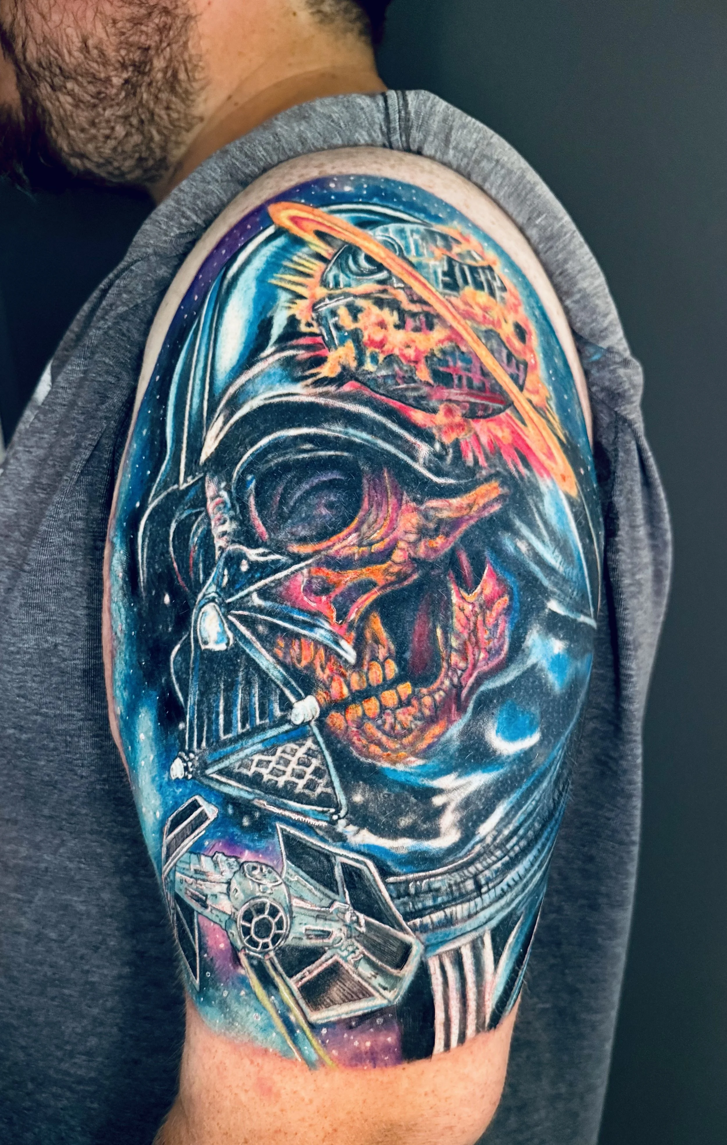 A colorful fantasy space-themed tattoo on a person's upper arm featuring a skull with a galaxy background, a spaceship, a planet, and various space elements.