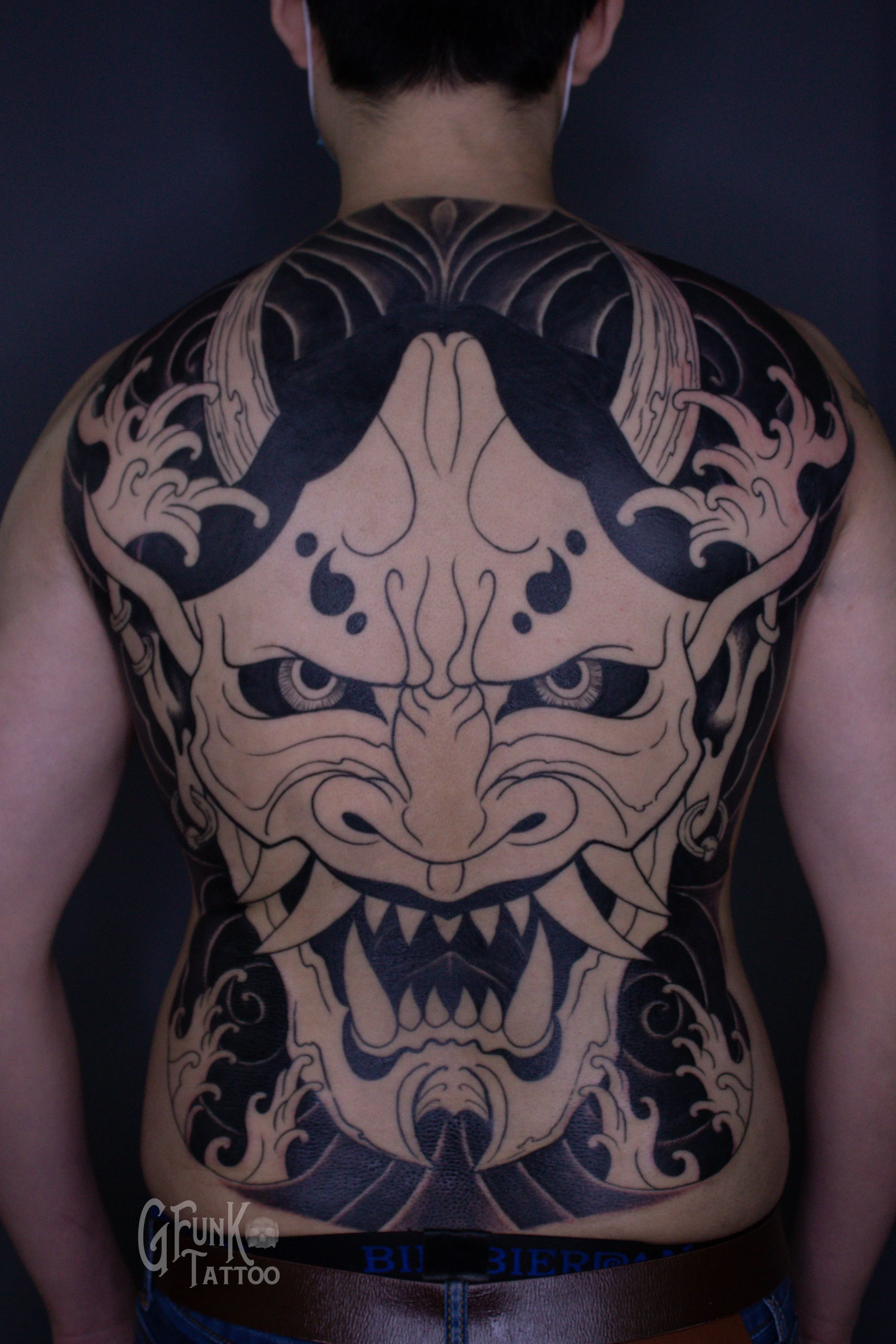 Large black and gray tribal-style tattoo of a fierce face with sharp teeth and intense eyes covering the person's chest and shoulders.