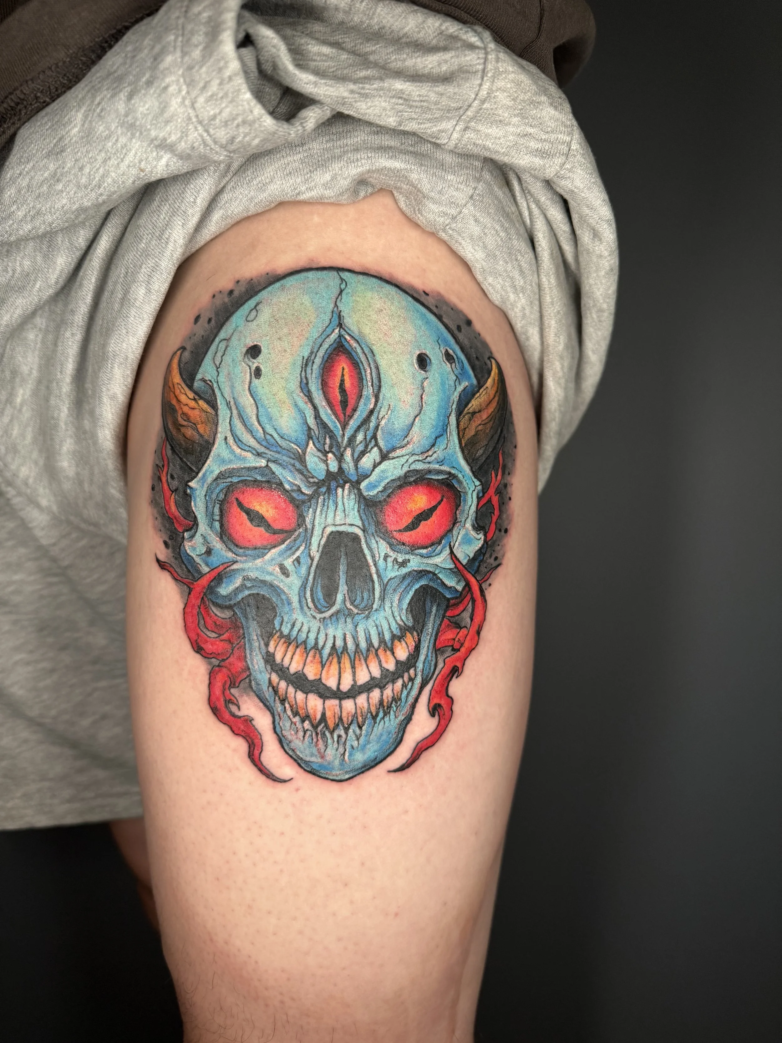 Colorful tattoo of a skull with glowing red eyes, blue and green hues, orange horns, and red flames around the face on a person's upper arm.