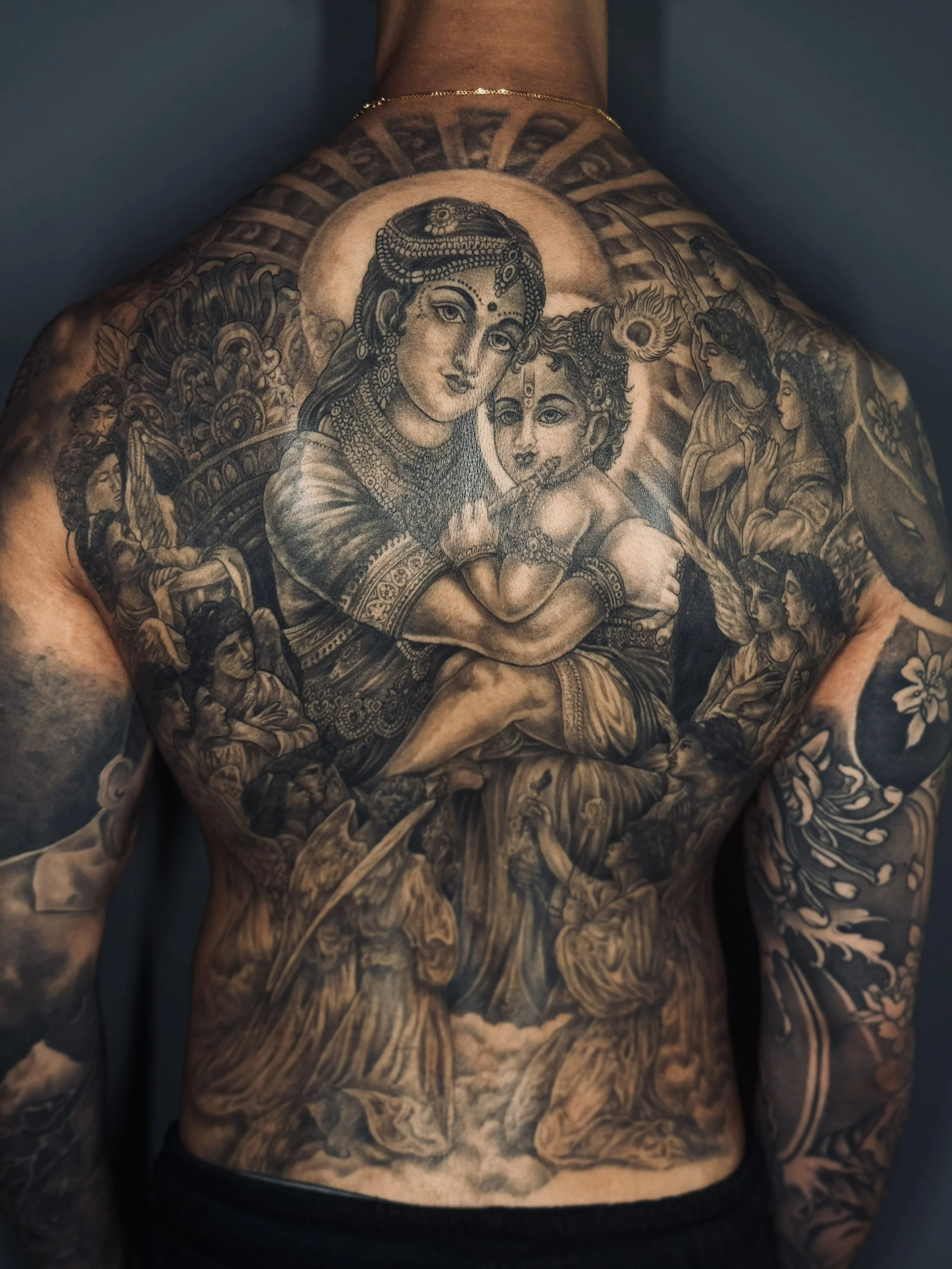 A detailed black and grey tattoo depicting a woman with jewelry and a child on her lap, surrounded by smaller figures, floral patterns, and decorative elements covering the chest and arms.