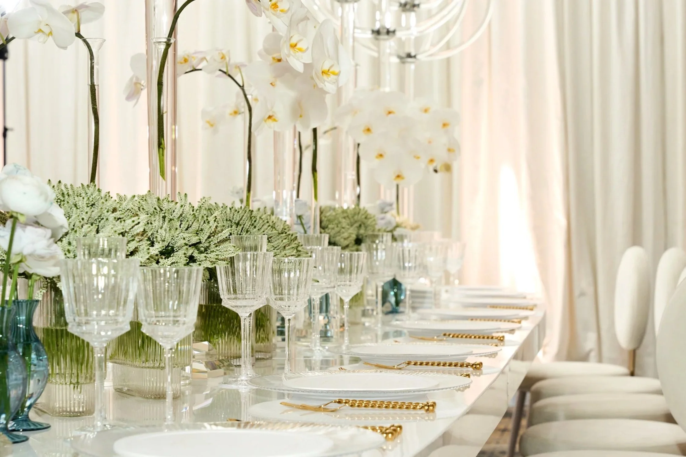 Elegant table setting with white plates, gold utensils, crystal glasses, and white floral centerpieces, set in a bright room with cream-colored curtains.