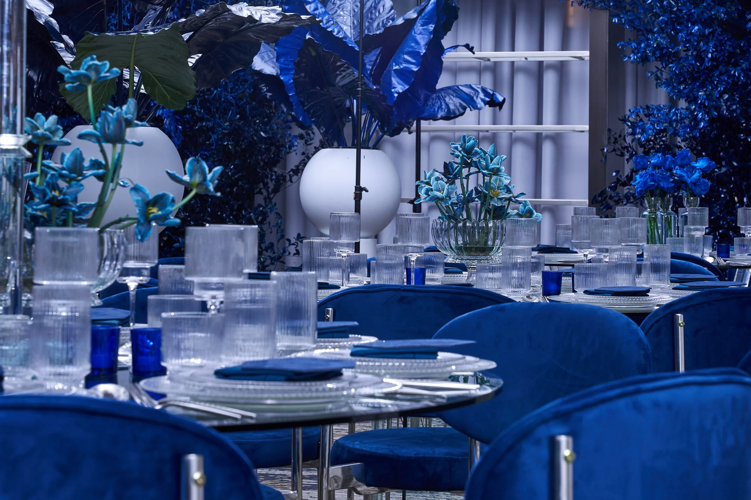 A dining setup with tables and chairs decorated in shades of blue, featuring glassware, napkins, and floral centerpieces with blue flowers and large blue leaves.