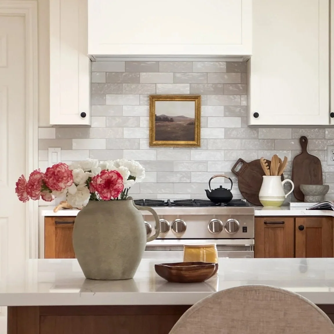 A kitchen with white cabinets, a gray and white tiled backsplash, a stove with four knobs, a black kettle, wooden cutting boards, a white pitcher with wooden utensils, a gray bowl, a book, a beige vase with pink and white flowers, and a wooden bowl on a white countertop.