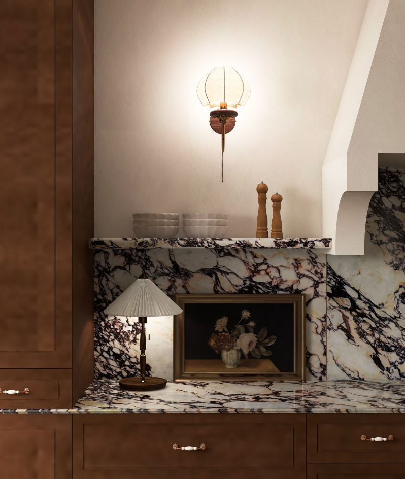 Close-up of a kitchen countertop with a marble pattern, a small table lamp with a pleated shade, a framed painting of flowers, and two wooden salt and pepper shakers, with a marble backsplash and upper shelf holding bowls and a wall-mounted light fixture.