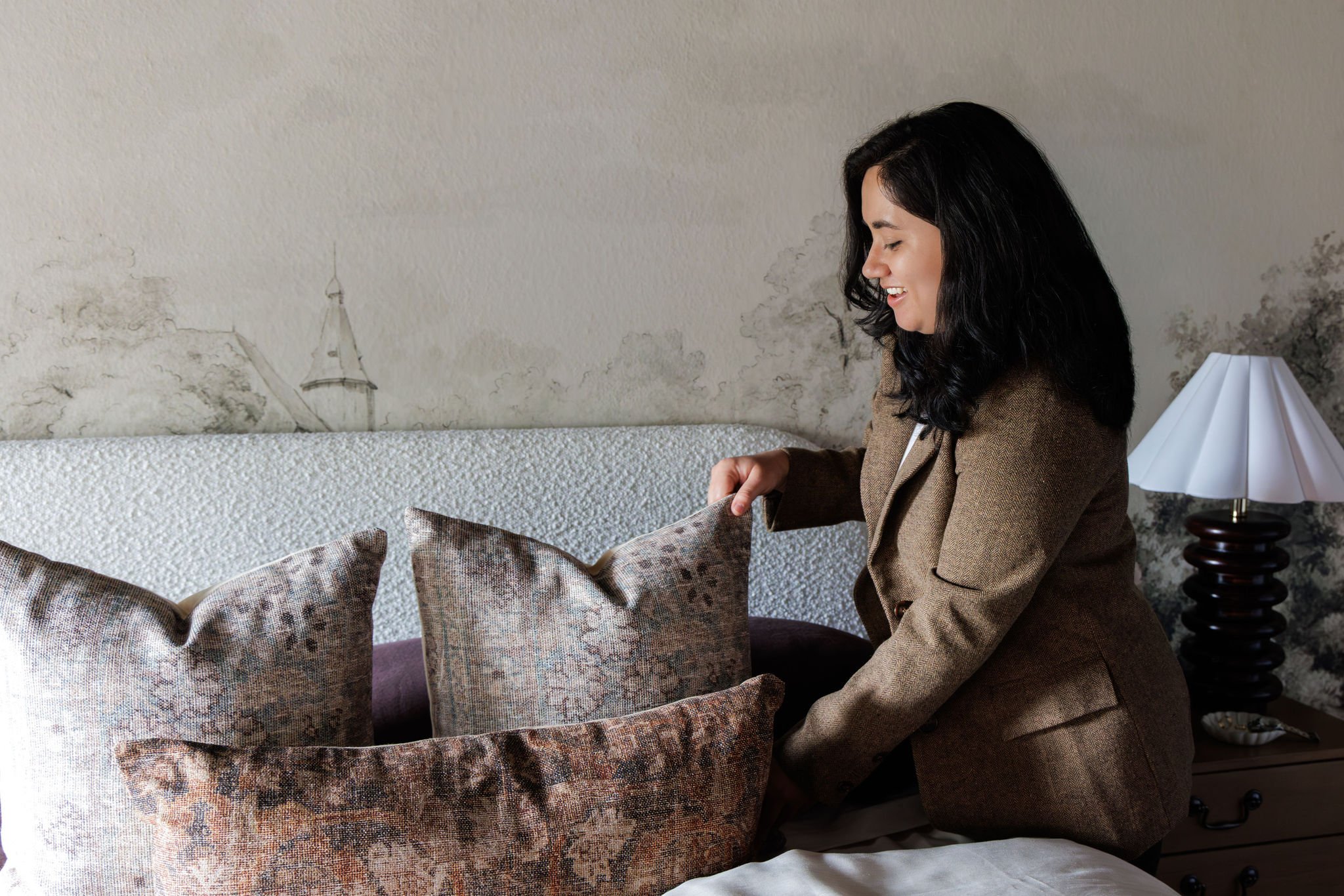 A woman with black wavy hair smiling and sitting on a bed with several patterned pillows, in a room with a textured wall, a white lampshade on a dark wooden lamp, and a light gray bedspread.