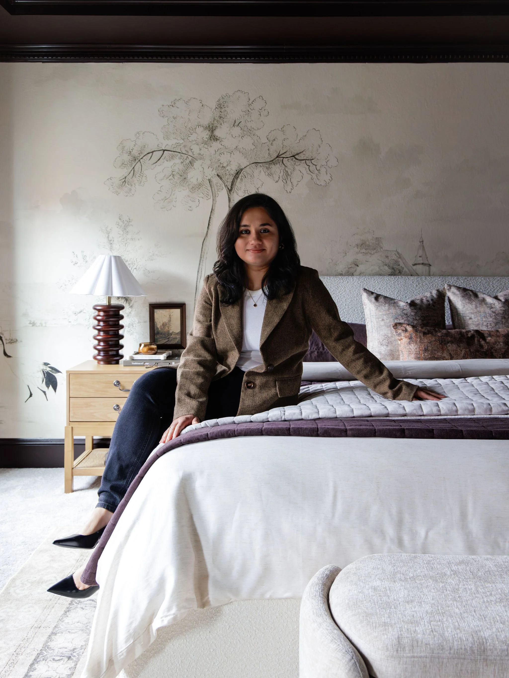 A woman sitting on a bed with a dark brown blazer and black heels, smiling at the camera in a bedroom with a large mural of a tree and a sailboat on the wall behind her, a lamp, a framed picture, and decorative pillows on the bed.