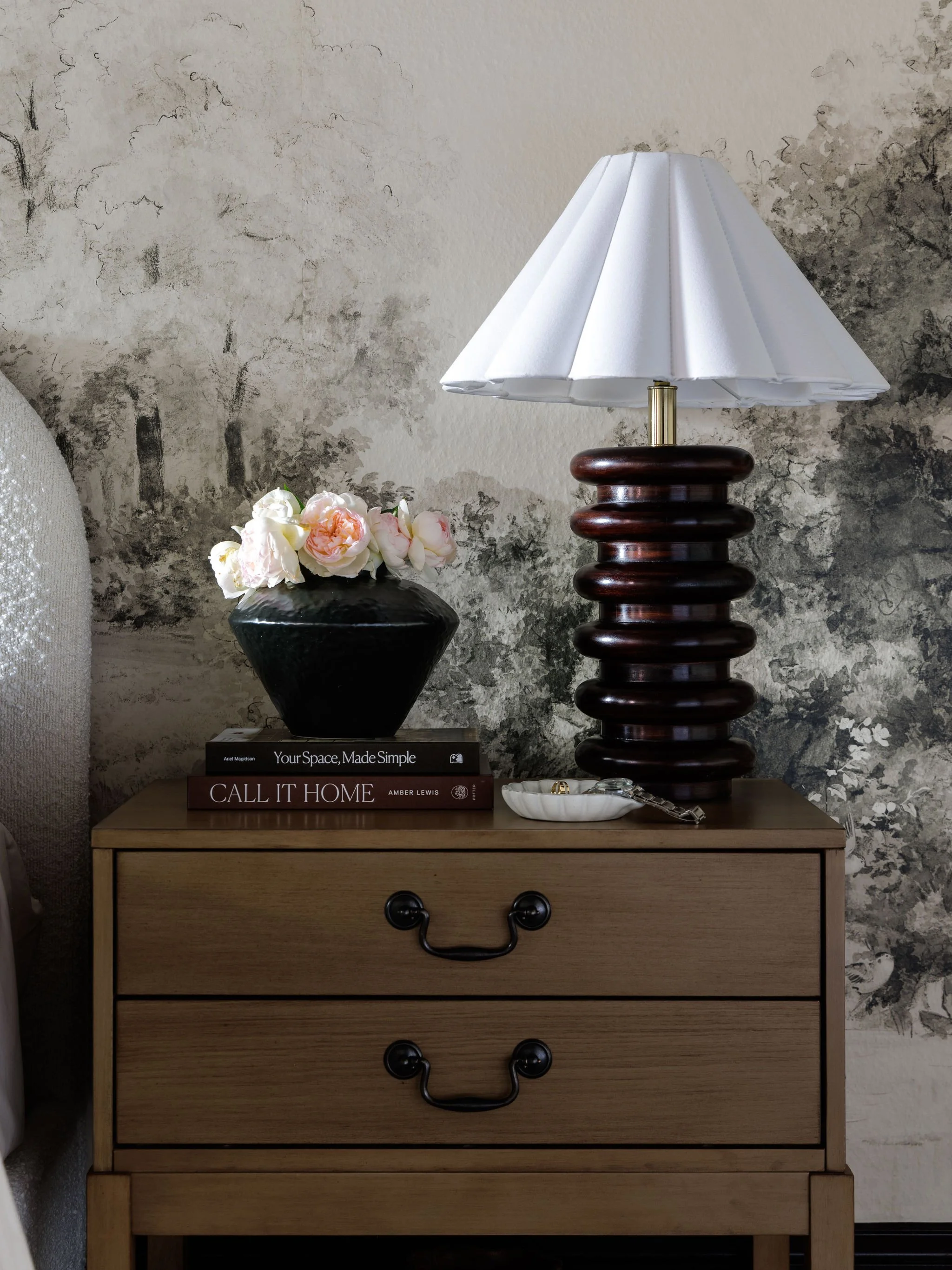 A wooden dresser with three drawers, black metal handles, a lamp with a dark wooden base and white lampshade, a black vase with flowers, three books, and a small dish with jewelry on top, against a textured wall with artwork.