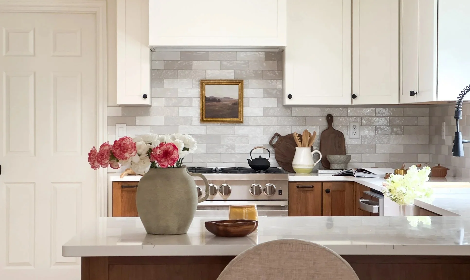 Kitchen with white upper cabinets, wooden lower cabinets, and a gray tile backsplash. There are flowers on the counter and a large ceramic vase with pink and white flowers in the foreground. A small black teapot sits on the stove, with wooden cutting boards, a bowl, and an open book behind it. A framed landscape painting hangs on the wall above the stove.