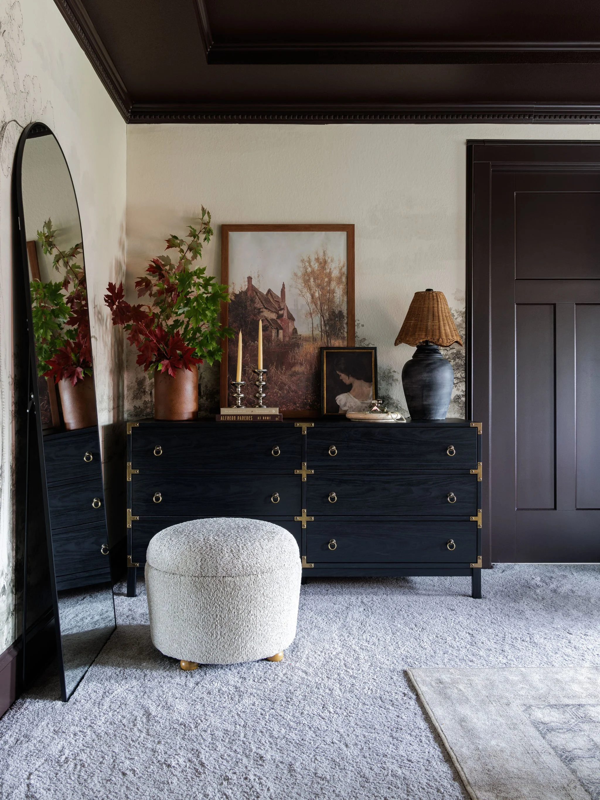 A modern interior room featuring a black wooden dresser with gold accents, decorated with framed artwork, tall candlesticks, a porcelain vase with branches, a ceramic table lamp, and a small beige ottoman, with a mirror on the left side and a dark wood-paneled wall on the right, on a plush light-colored carpet.