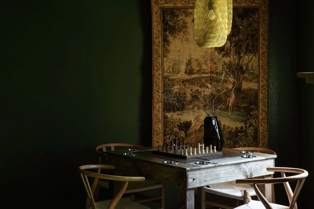 A cozy dining area with a square wooden table, four chairs, a black vase with thin branches, a chessboard, and a yellow woven pendant light hanging above. Behind, a large tapestry depicting a nature scene with trees and a river.
