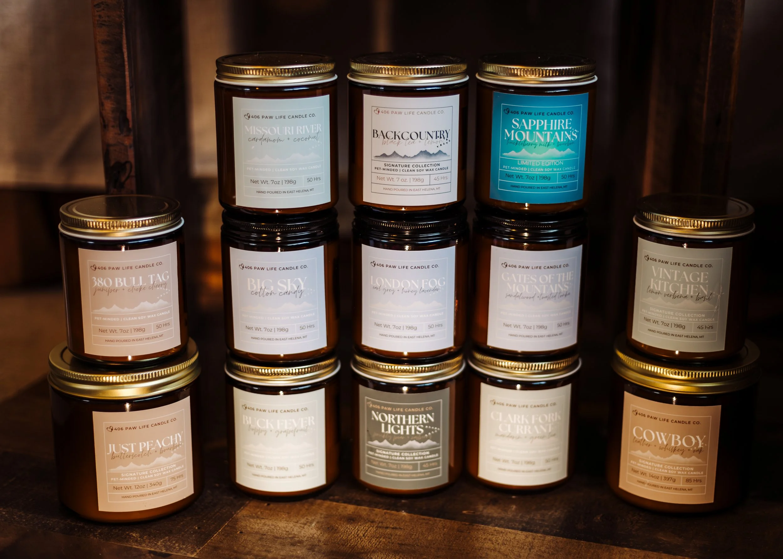 A display of 13 brown glass candles with gold lids, arranged on a wooden shelf. The candles have white labels with different names and scents, including Missouri River, Backcountry, Sapphire Mountains, 380 Bull Tag, Big Sky, London Fog, Gates of the Mountains, Vintage Kitchen, Just Peachy, Buck Fever, Northern Lights, Clark Fork River, and Cowboy.