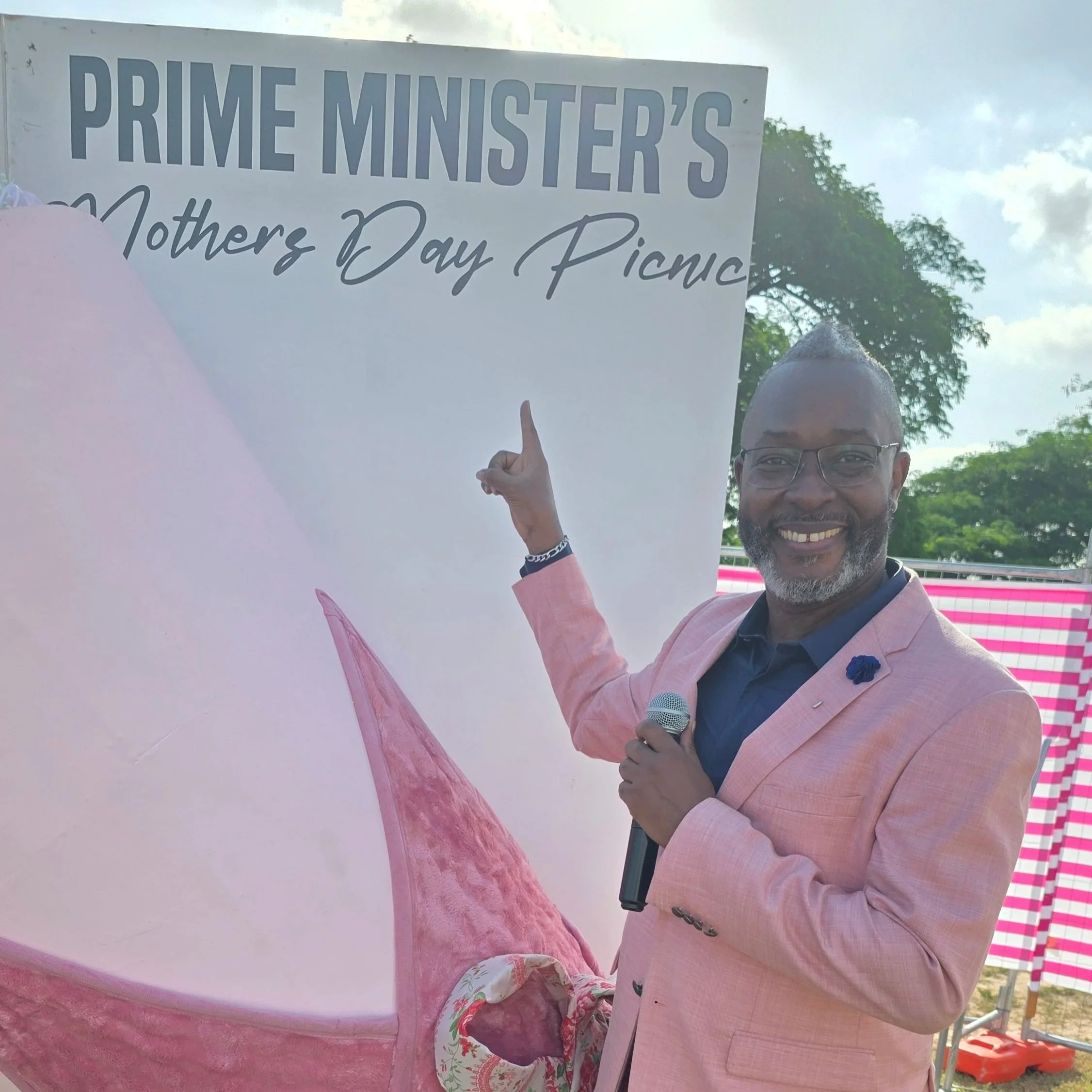 A man wearing glasses and a pink blazer is smiling and holding a sign that reads, "Prime Minister's Mother's Day Picnic." He is outdoors, with trees, a barrier, and a partly cloudy sky in the background.