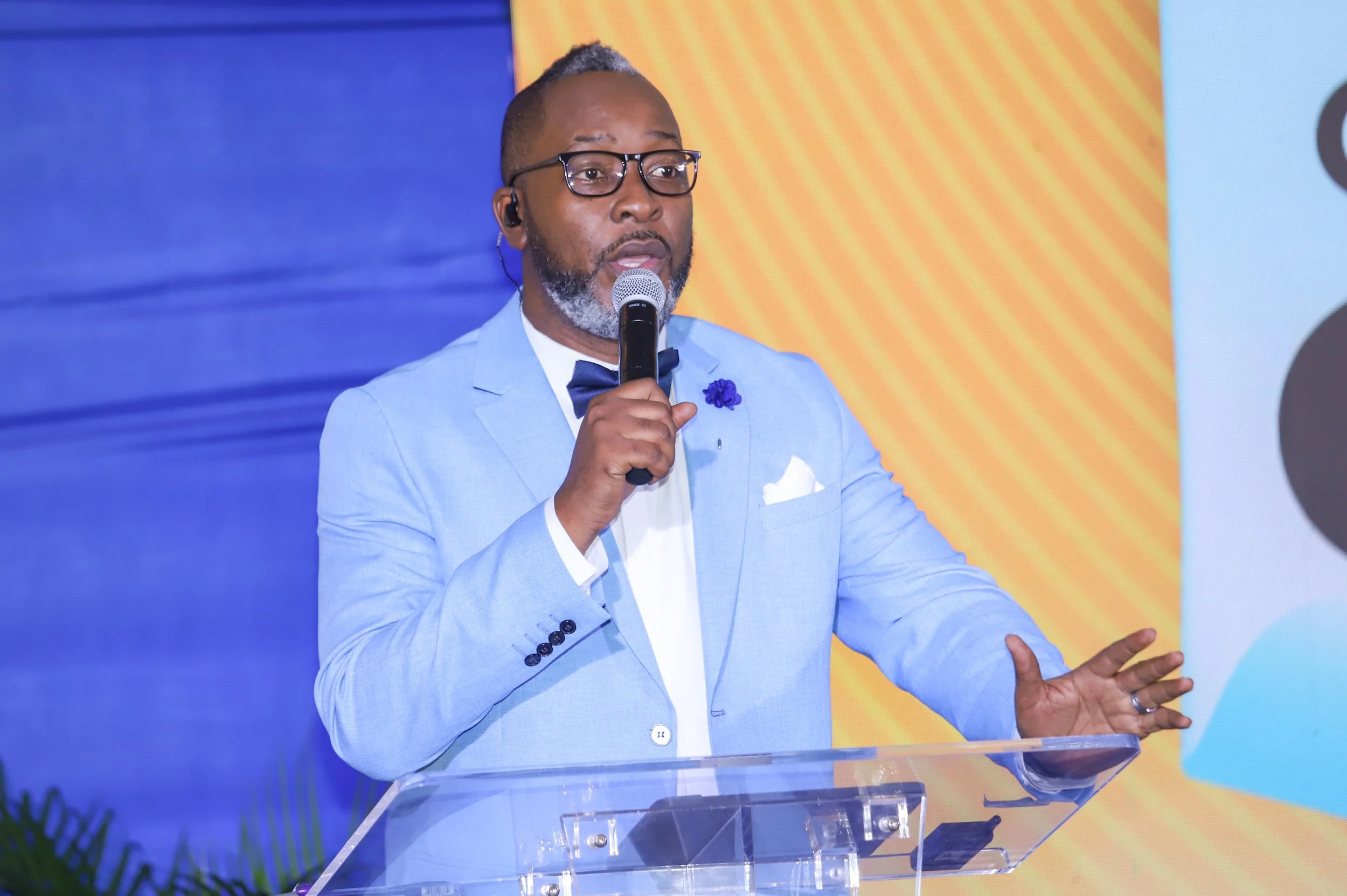 A man in a light blue suit, white shirt, and glasses speaking into a microphone at a podium against a colorful background.