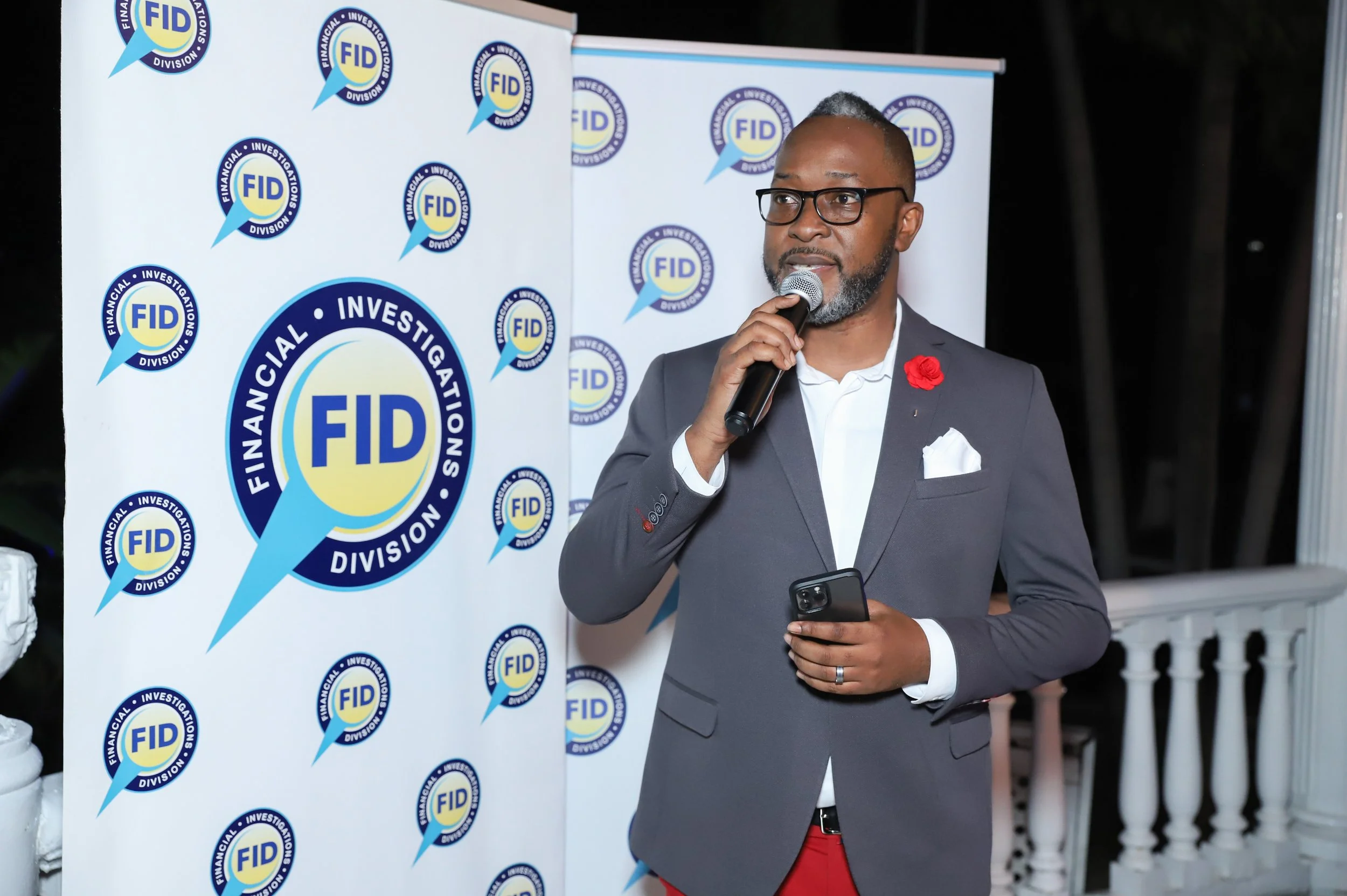 A man with glasses and a beard, wearing a dark blazer and white shirt, holds a microphone and stands in front of a backdrop with multiple FID logos, speaking at an event.