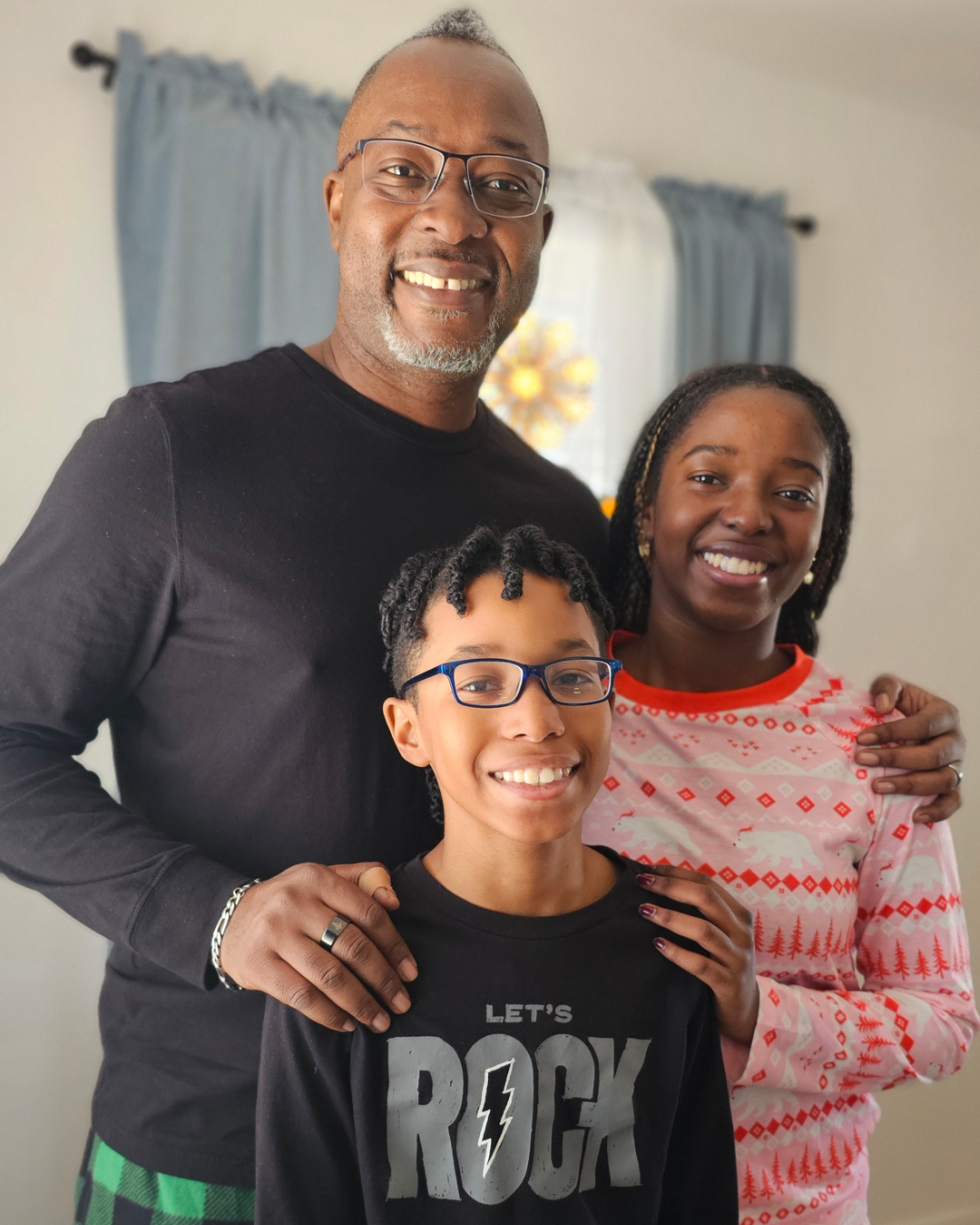 A smiling family of three standing together indoors. The father, wearing glasses and a black shirt, stands behind the children with his hand on one child's shoulder. The girl, with braided hair and wearing a festive pink and white sweater, is on the right. The boy, with glasses and short curly hair, is in front wearing a black T-shirt that says 'Let's Rock' with a lightning bolt.