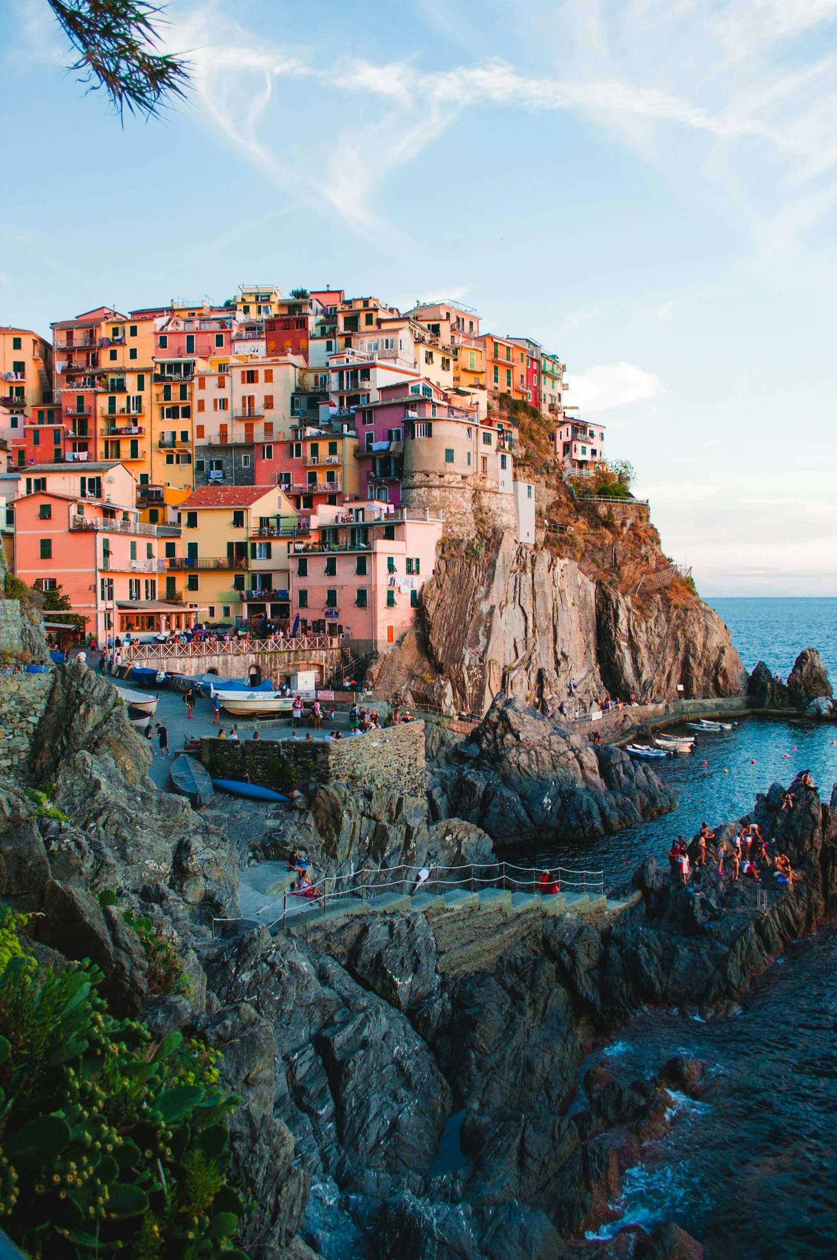 Colorful houses built on a cliff by the sea in Cinque Terre, Italy, with boats in the water and people on the rocky shoreline during sunset.