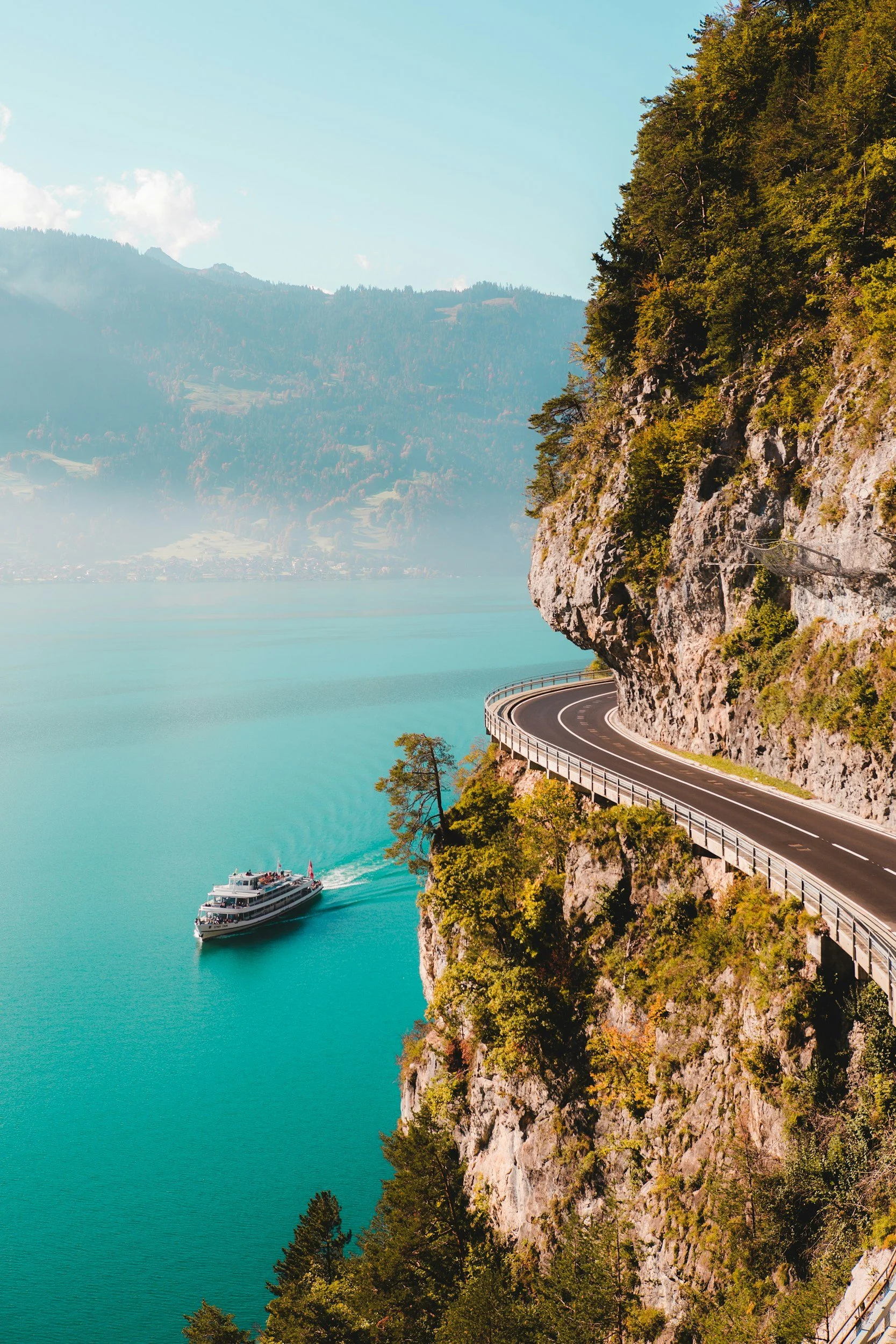 A winding road built into a rocky cliffside above a turquoise lake, with a boat sailing on the water and mountain scenery in the background.