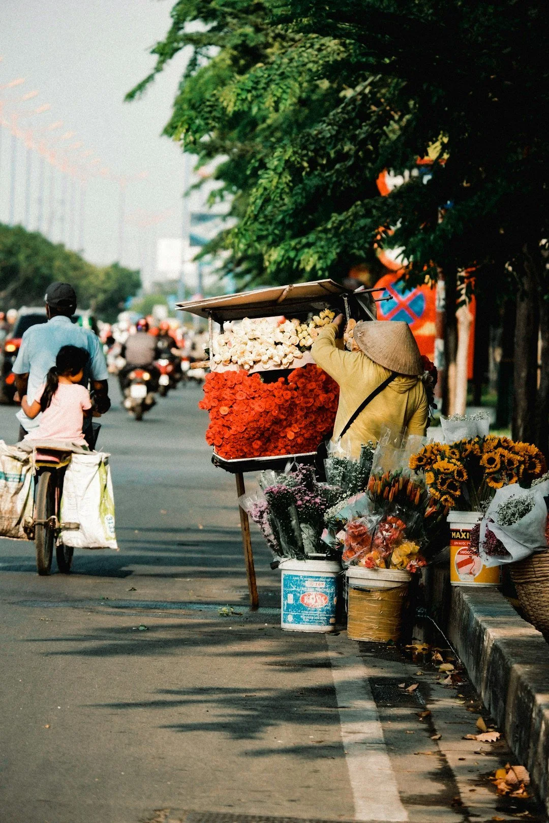 Street vendor selling flowers on the sidewalk with people walking and riding bikes on the busy street.