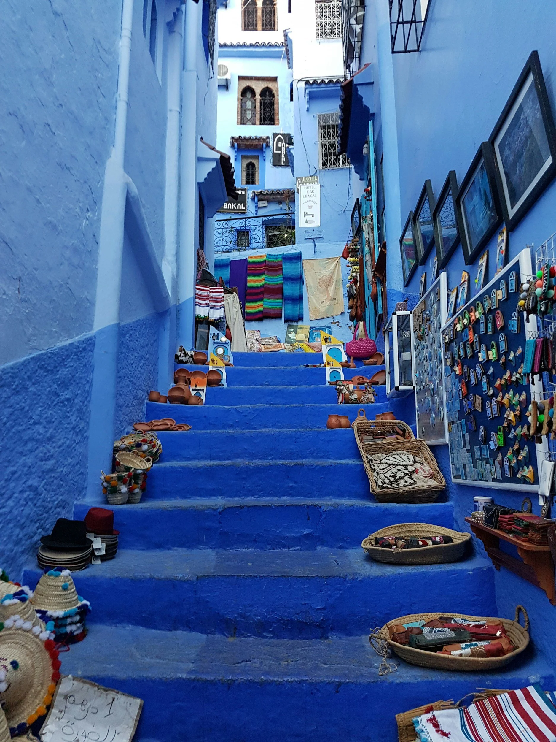 Colorful marketplace on blue stairs and walls in Chefchaouen, Morocco, with various souvenirs and textiles for sale.