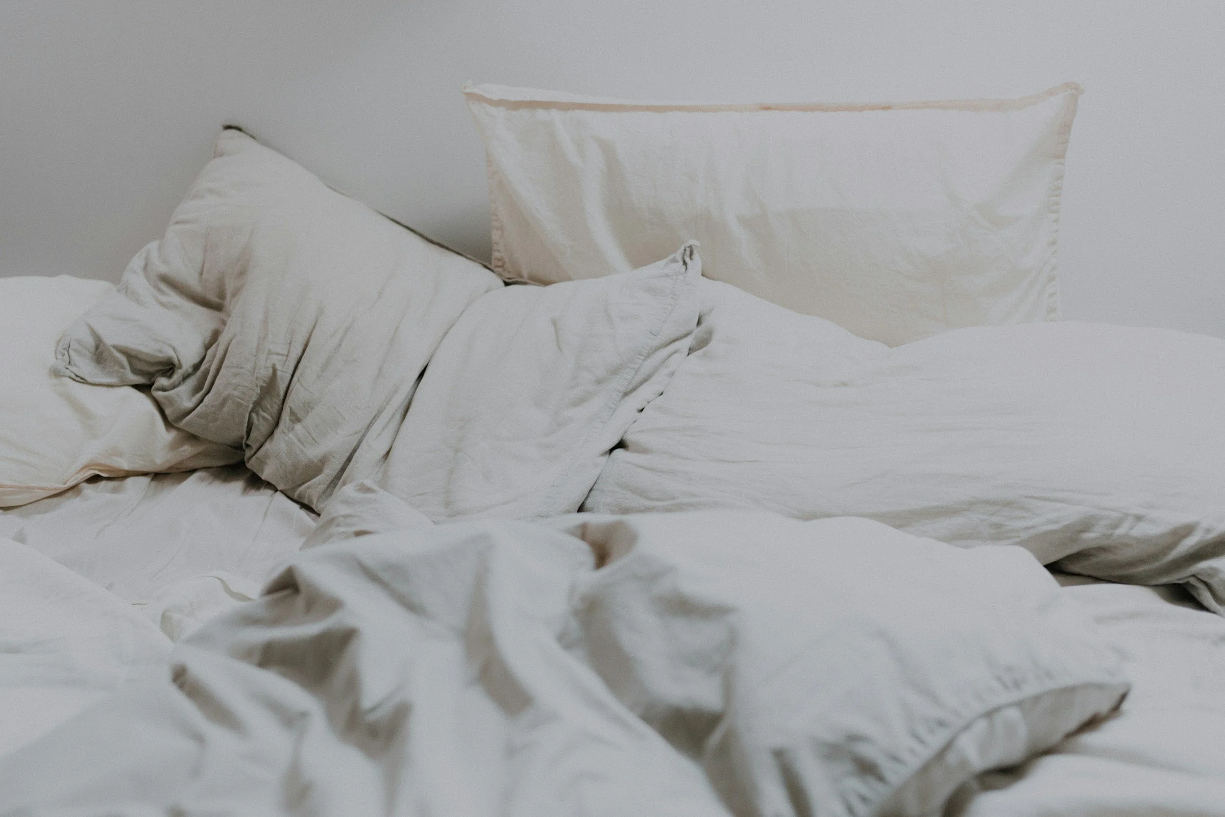 Disheveled white bed with wrinkled sheets and multiple pillows, some leaning against a plain wall.