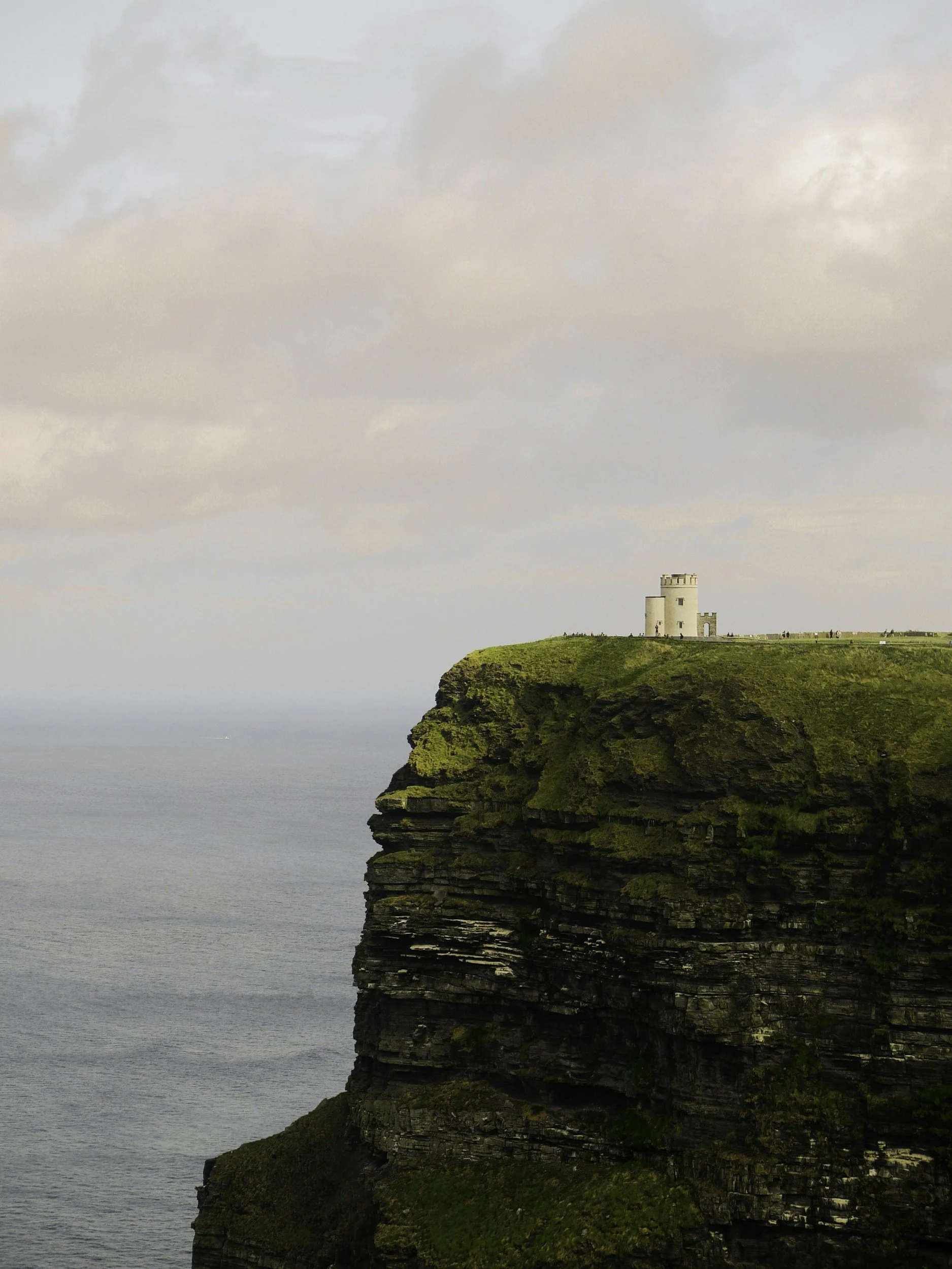 A cliff with lush green grass and layered rock formations, with a white tower structure on top, overlooking the ocean under a cloudy sky.