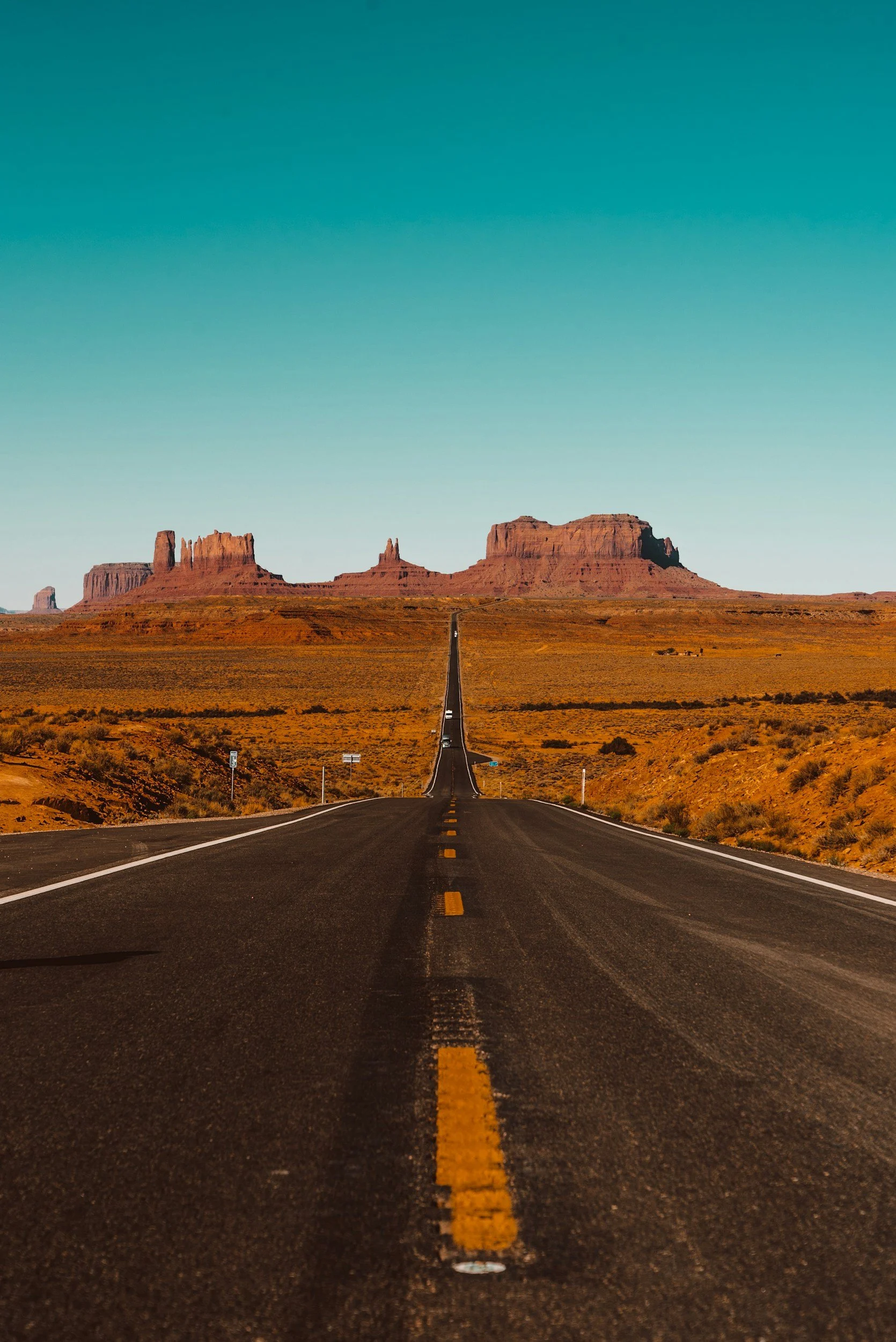 Long straight road leading through desert landscape with iconic red rock formations in the distance under a clear blue sky.