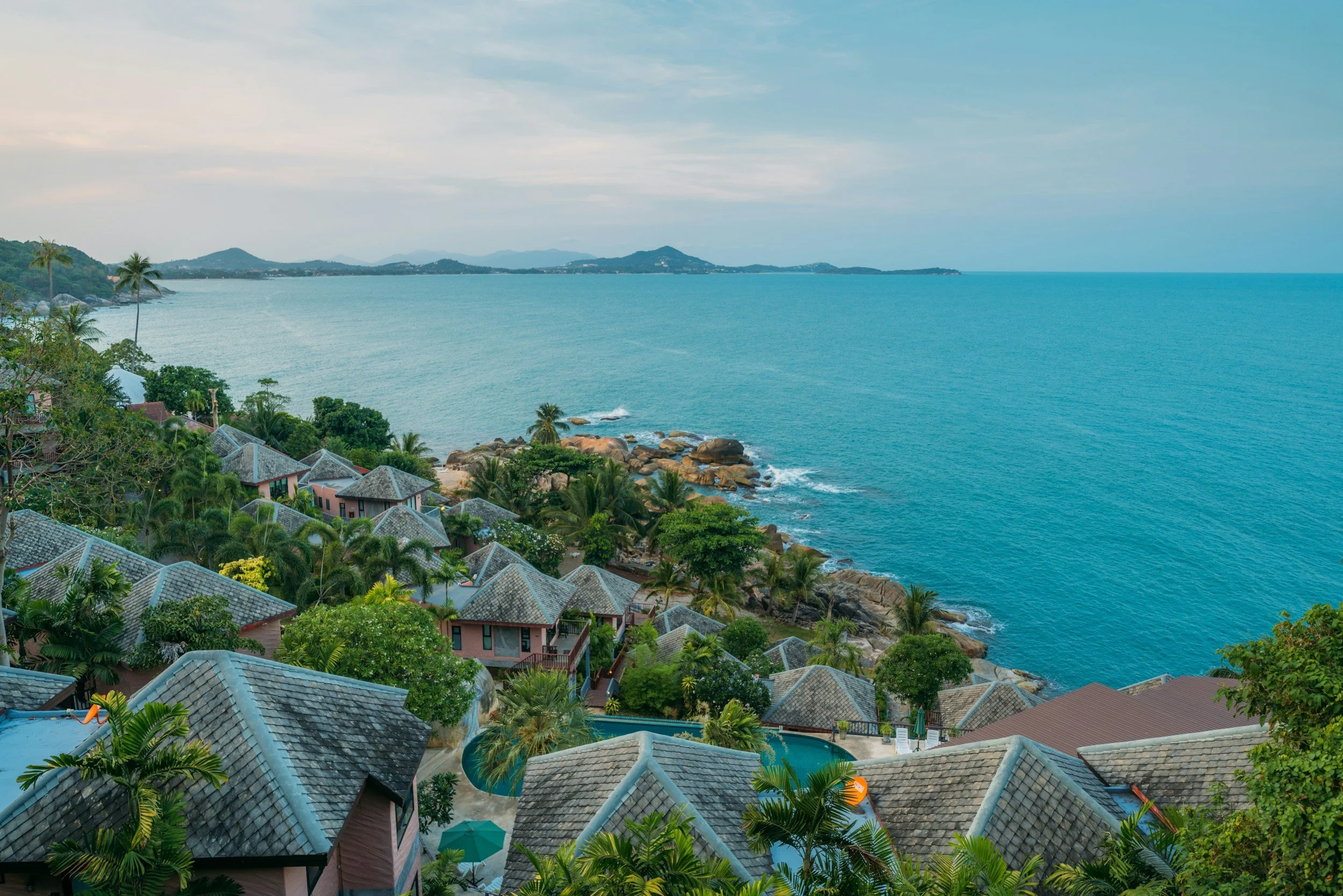 Coastal view of a resort with multiple bungalows surrounded by lush greenery, palm trees, and a swimming pool. The ocean extends towards distant hills, with rocky shoreline and cloudy sky.