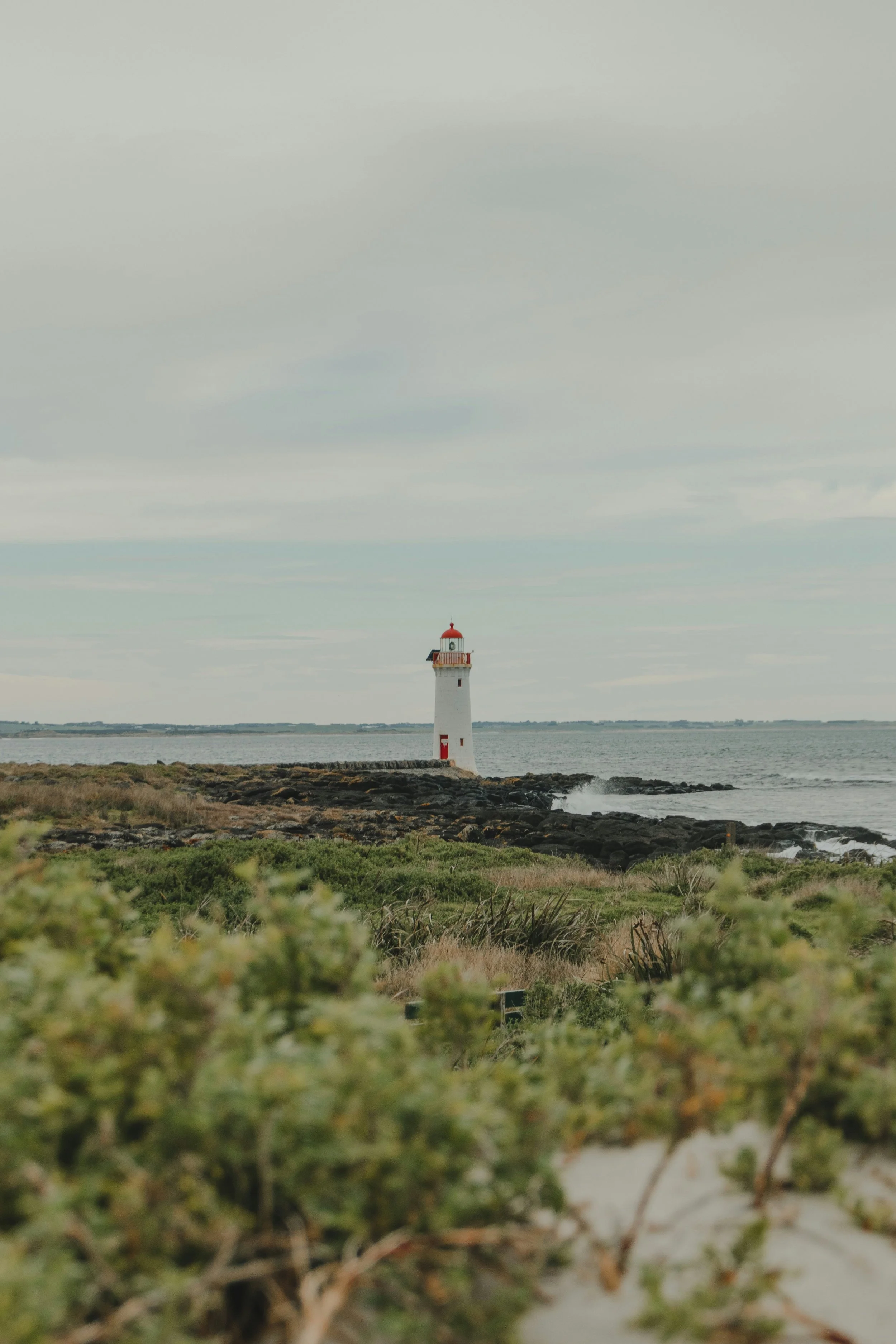 Lighthouse on rocky coast with green shrubs and cloudy sky in the background.